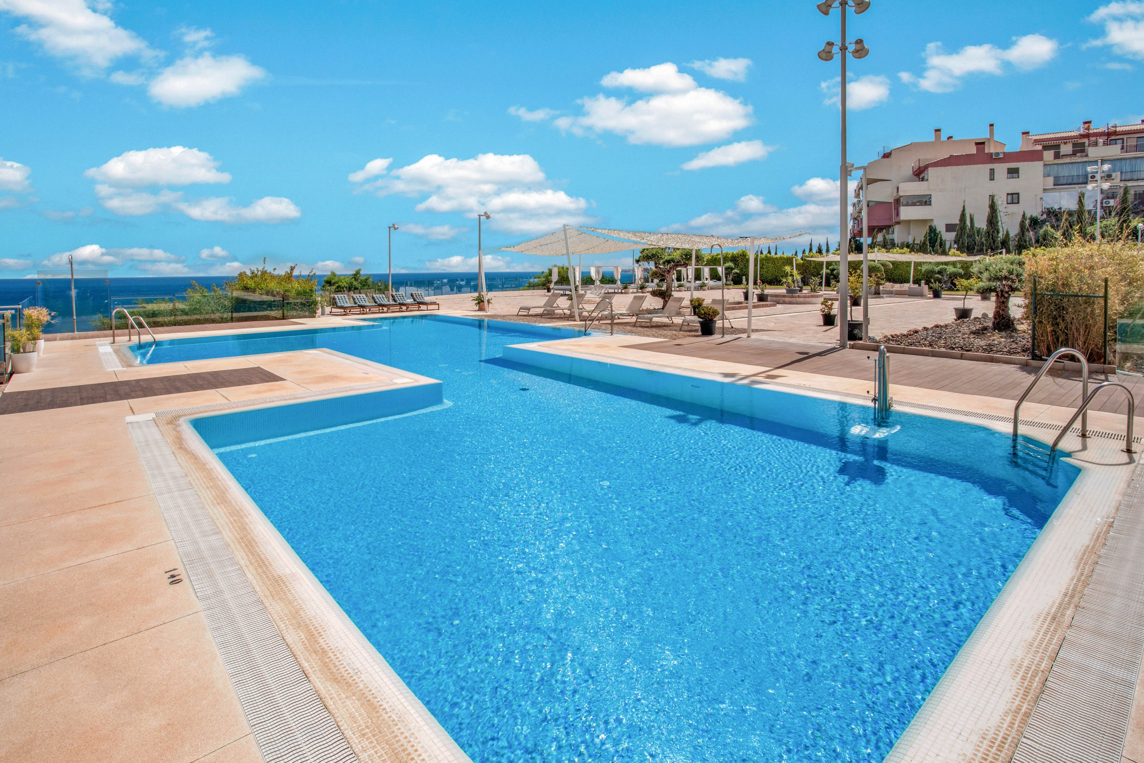 A bright blue swimming pool stretches across an outdoor terrace under a clear sky with scattered clouds. Lounge chairs and umbrellas line the poolside area, suggesting a relaxing resort or hotel setting. Metal ladders and safety railings frame the pool, while landscaped greenery and buildings appear in the background. The distant ocean horizon enhances the open, tranquil atmosphere.