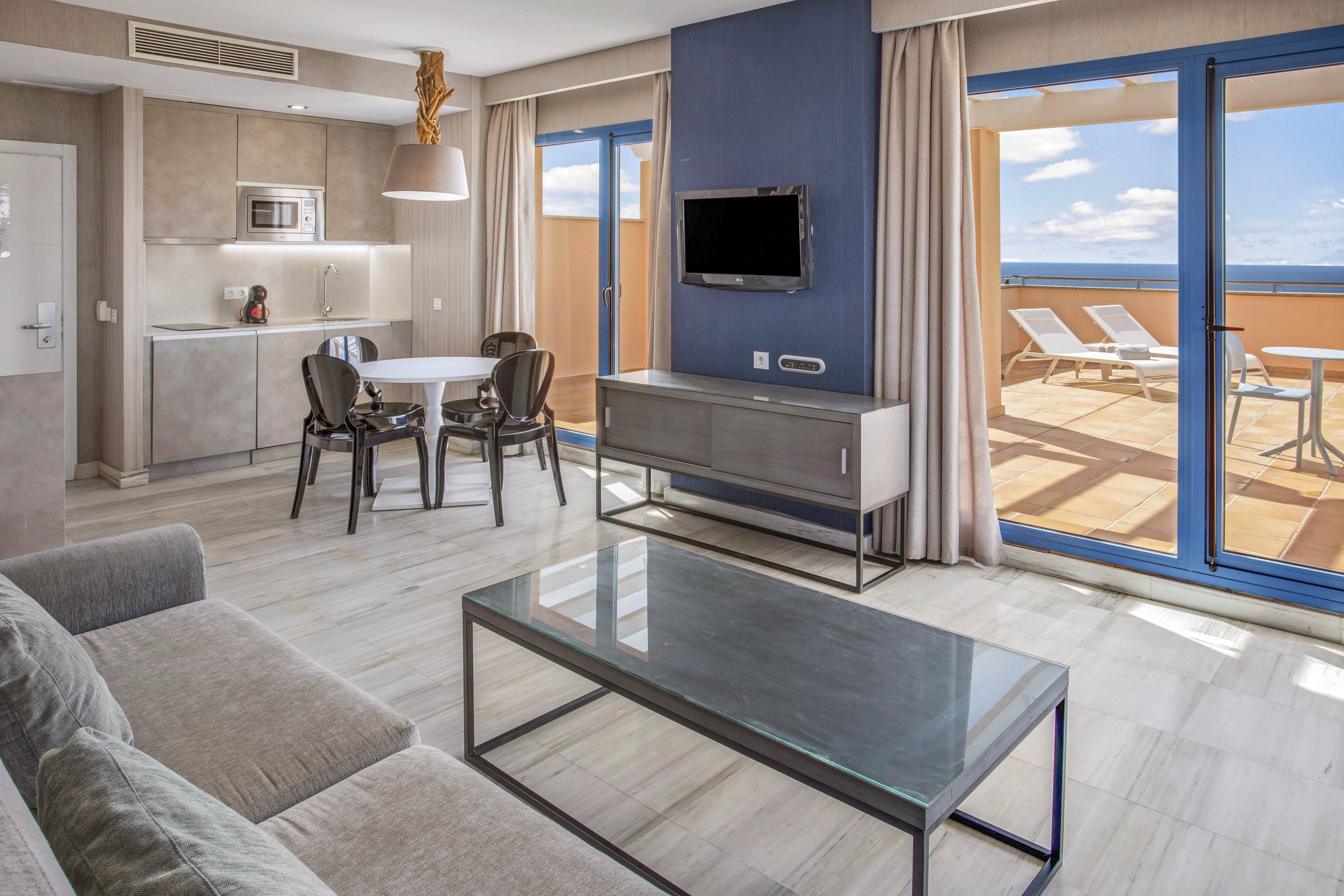 A contemporary hotel suite features a cozy living area with a gray sofa and glass coffee table. A compact kitchenette and round dining table with dark chairs sit near the entrance. Large sliding glass doors open to a spacious sunlit terrace with lounge chairs and an ocean view. Neutral tones, clean lines, and blue accents create a bright, minimalist atmosphere.