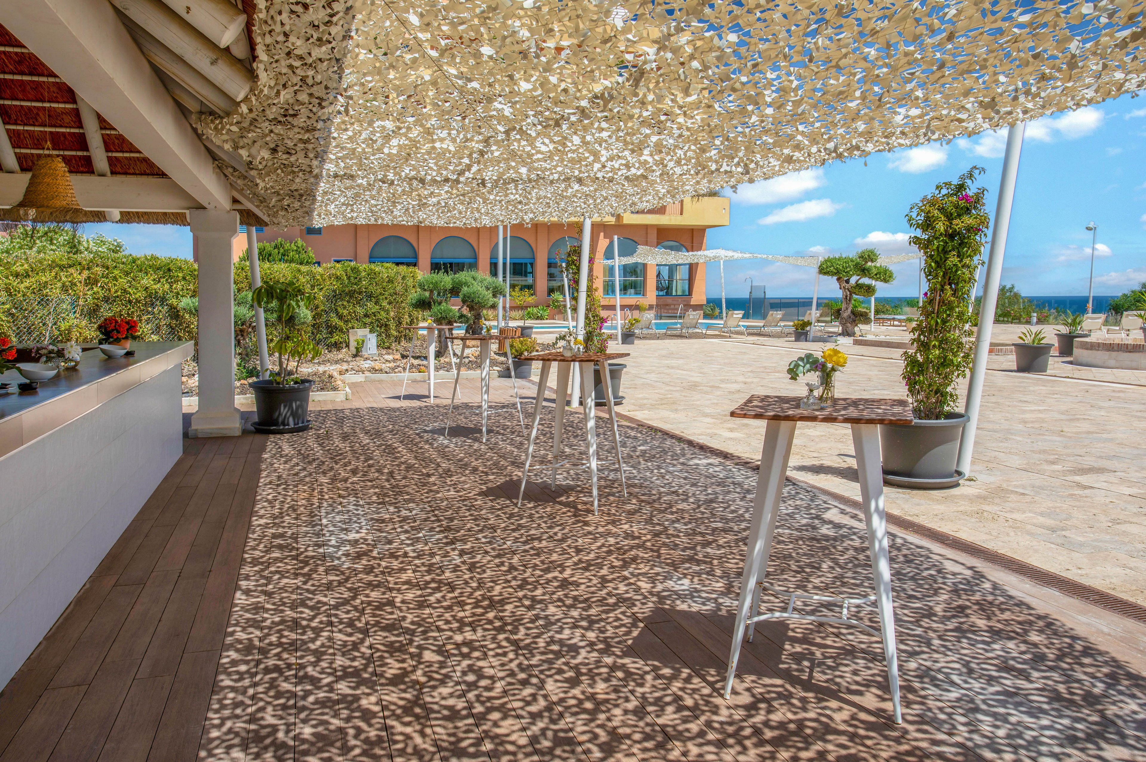 An outdoor seaside terrace features a shaded bar area with high tables and stools arranged along a wooden deck. A camouflage-style canopy casts intricate shadows on the floor, creating a dappled light effect. In the background, a large building with arches faces an open plaza and the ocean beyond. Potted plants and small floral arrangements on the tables add greenery and a relaxed, summery atmosphere.