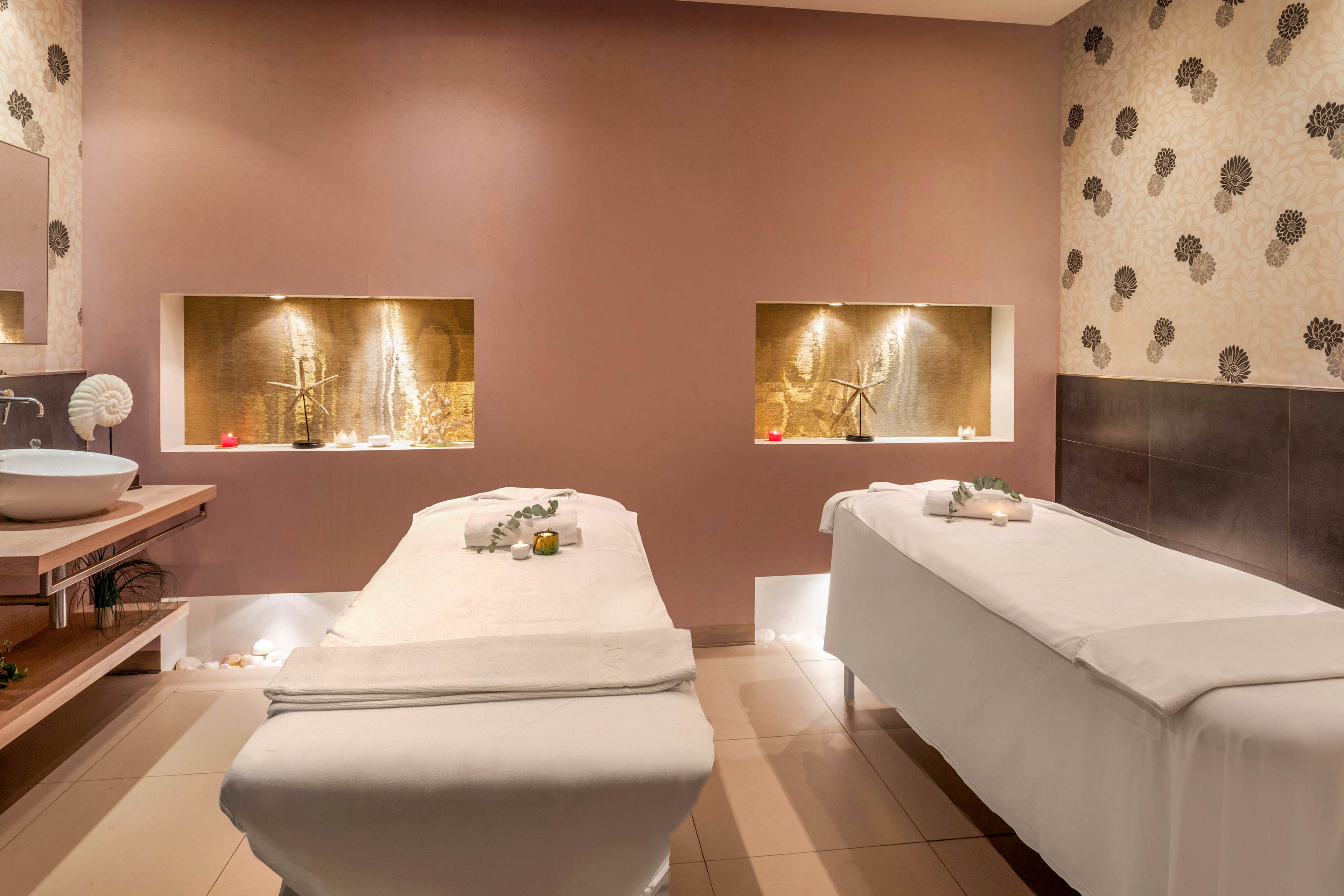 A serene spa treatment room features two neatly prepared massage tables facing a warm-toned wall. Built-in illuminated niches with decorative elements create a soft, ambient glow. A modern sink and wooden counter with towels and candles add to the sense of relaxation. Neutral colors and subtle floral wallpaper contribute to a calm, luxurious atmosphere ideal for wellness treatments.