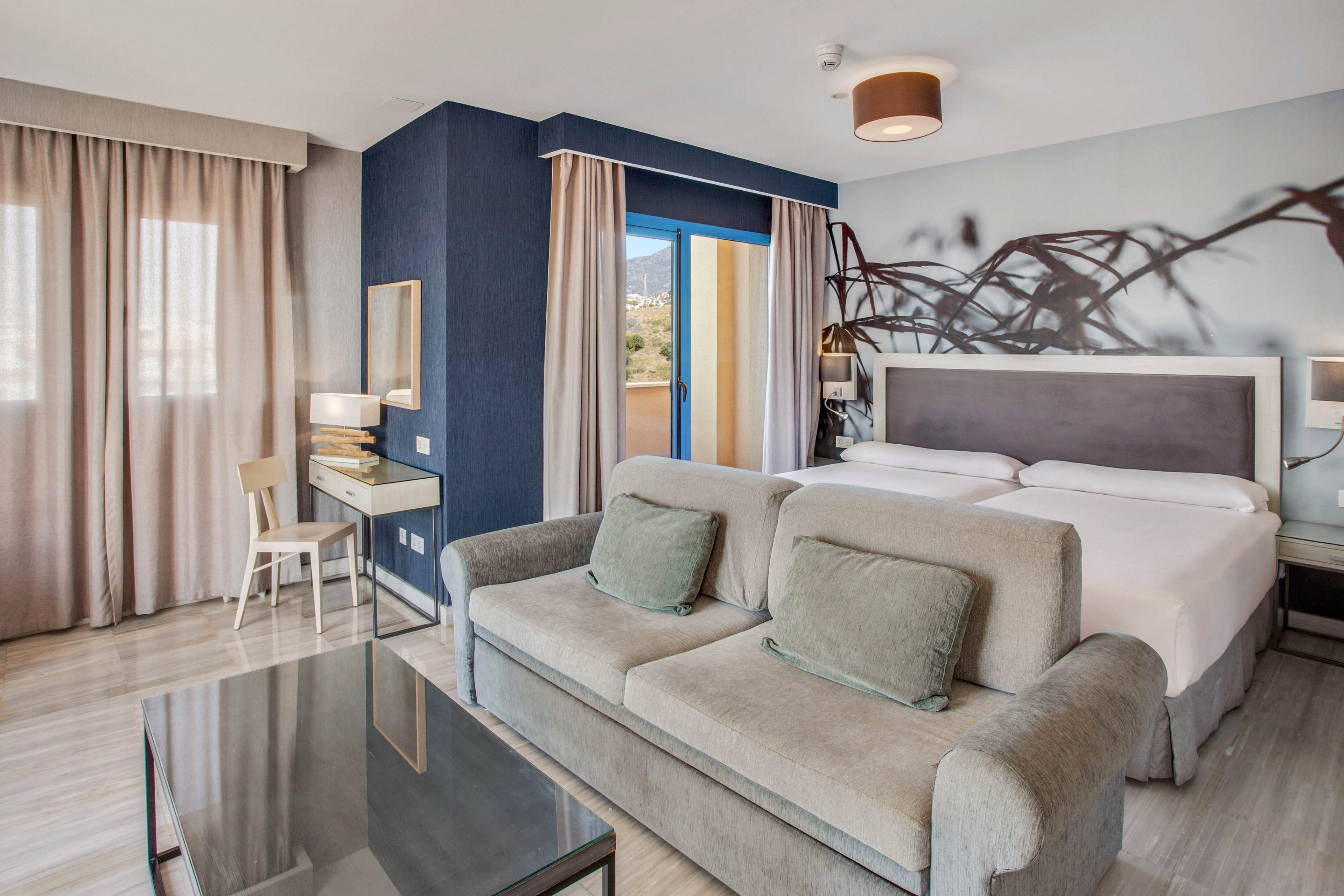 A contemporary hotel bedroom features a large bed with a padded headboard and neatly arranged white linens. A soft gray sofa with matching cushions faces a glass-topped coffee table in the foreground. Floor-to-ceiling curtains frame large windows and a balcony door, allowing natural light to brighten the room. A small desk and chair sit against a dark accent wall, while a subtle wall mural adds decorative interest.