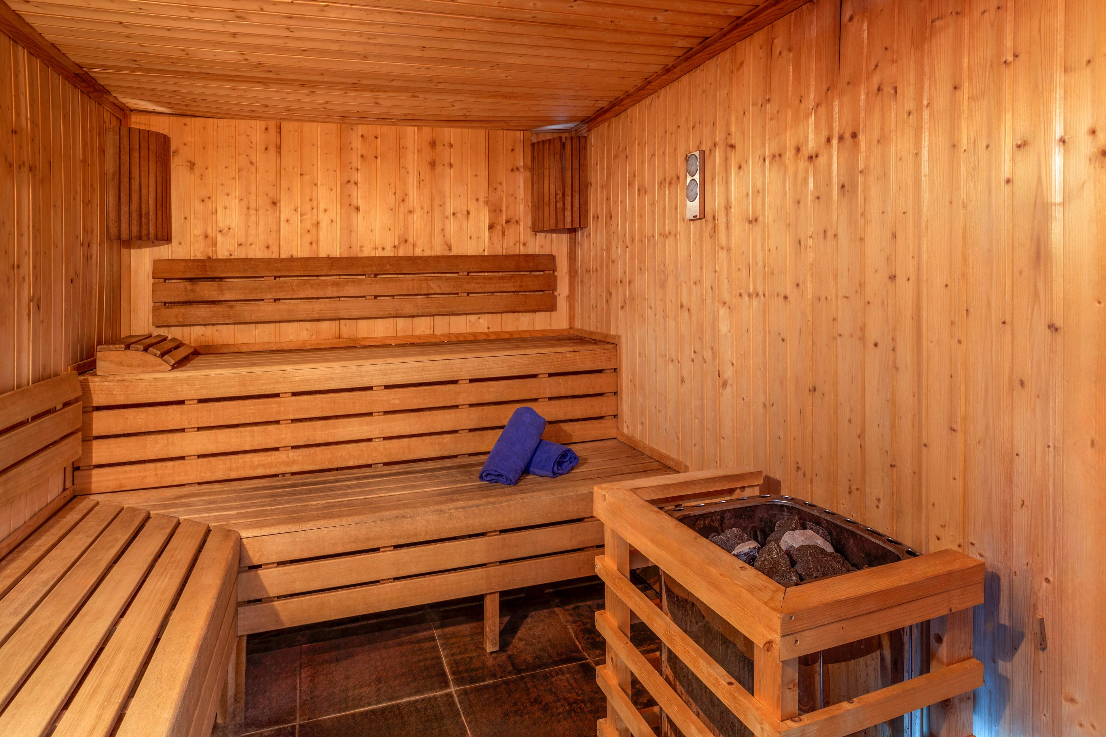 A cozy wooden sauna interior features tiered benches along the walls. Two neatly folded blue towels rest on the central bench, adding a pop of color to the warm wood tones. A metal heater filled with stones sits in the corner, ready to generate steam and heat. Soft lighting enhances the relaxing spa atmosphere and highlights the natural wood grain.