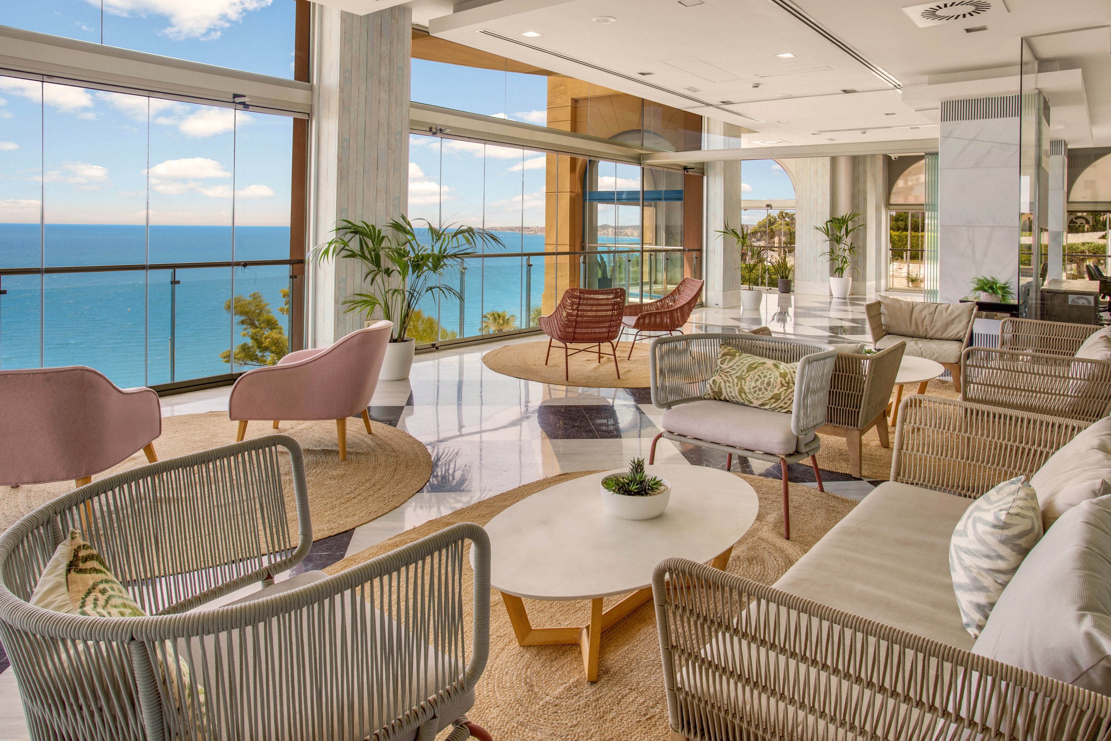 A contemporary hotel lounge features comfortable seating arranged around low coffee tables. Floor-to-ceiling windows reveal a bright blue sea and sky outside, creating an open, airy atmosphere. Neutral-toned sofas and woven chairs are accented with soft pink and green hues, while potted plants add a touch of nature. The space feels calm and inviting, ideal for relaxation or informal meetings with an ocean backdrop.