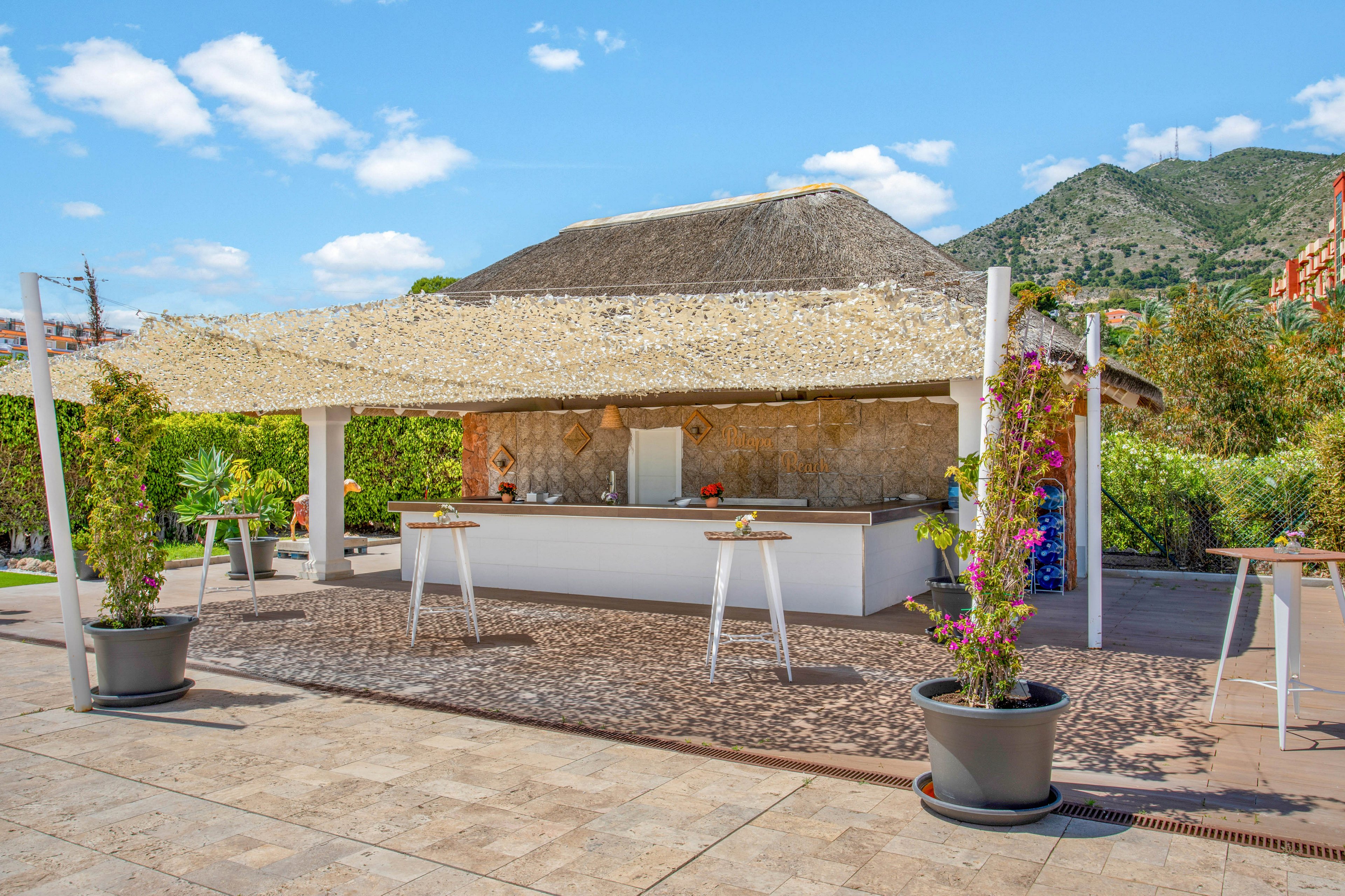 An open-air bar stands under a wide thatched canopy on a sunny day. Tall cocktail tables and potted plants decorate the tiled terrace in front of the counter. Green hedges and colorful climbing flowers frame the scene, adding a lush touch. In the background, mountains and scattered buildings rise under a bright blue sky with scattered clouds.