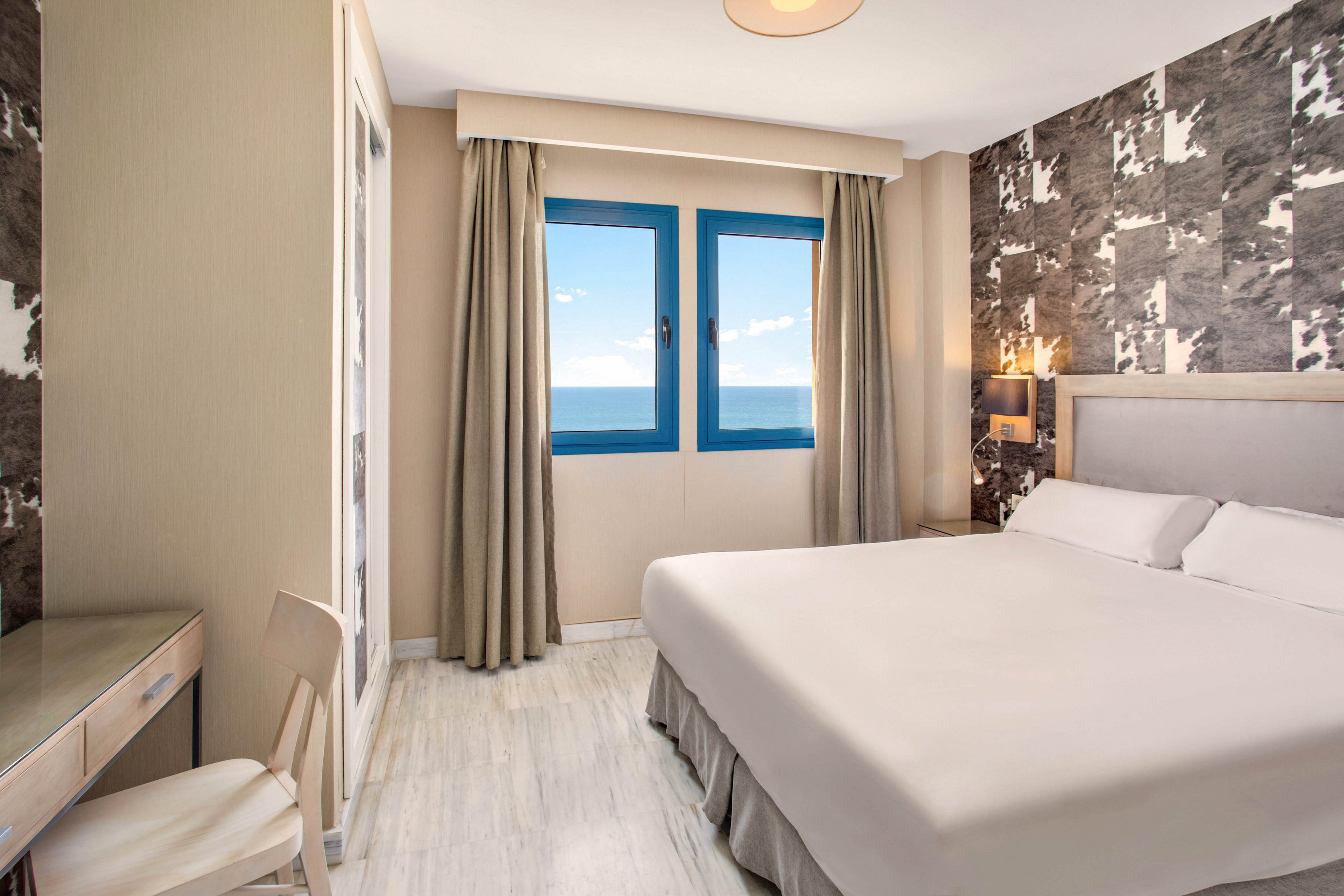 A contemporary hotel bedroom features a neatly made double bed with white linens. Large windows framed by beige curtains reveal a clear view of the blue ocean and sky outside. The room includes a small desk with a chair, a wardrobe with mirrored doors, and a patterned accent wall behind the bed. Neutral tones and clean lines create a calm, minimalist atmosphere ideal for relaxation.