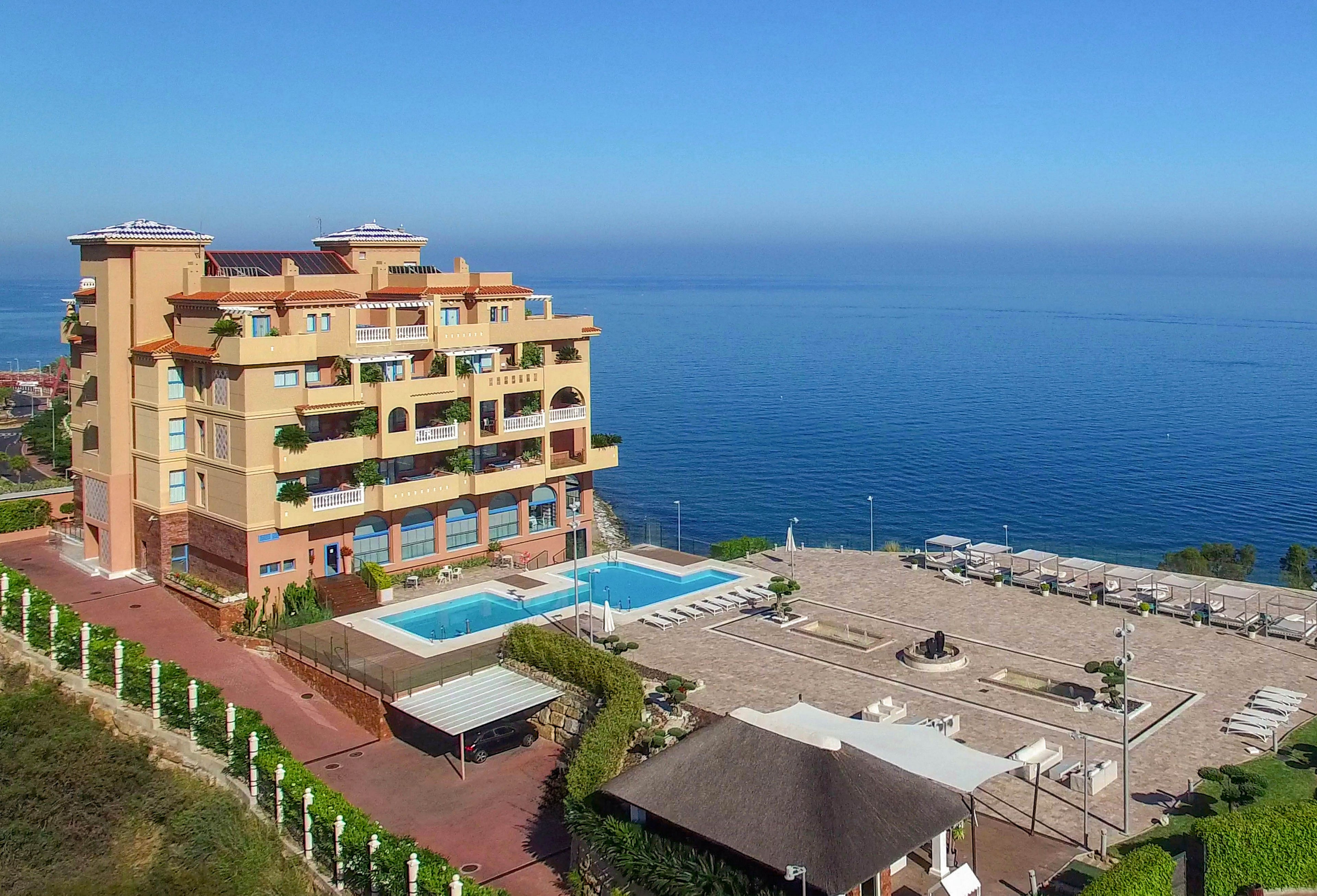 A multi-story hotel building stands beside a large swimming pool, overlooking a vast, calm ocean. The setting features a spacious terrace with sun loungers and shaded seating areas, ideal for relaxation. The scene is bright and clear, with a tranquil blue palette and lush greenery surrounding the property. No visible text or numbers are present in the image.