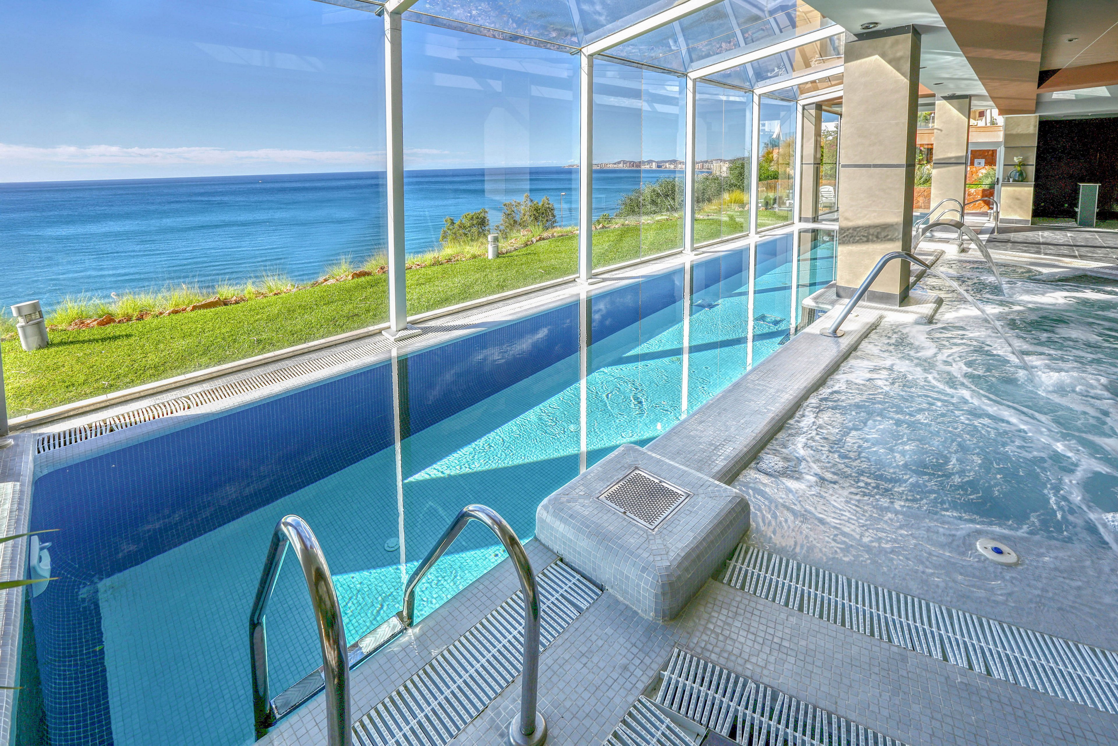 A modern indoor swimming pool is featured, enclosed by large glass panels that offer a panoramic view of the ocean and grassy landscape outside. The pool area includes stainless steel handrails and a bubbling spa section, creating a relaxing atmosphere. Natural light floods the space, highlighting the clean lines and tranquil blue palette. No visible text or numeric values are present in the image.