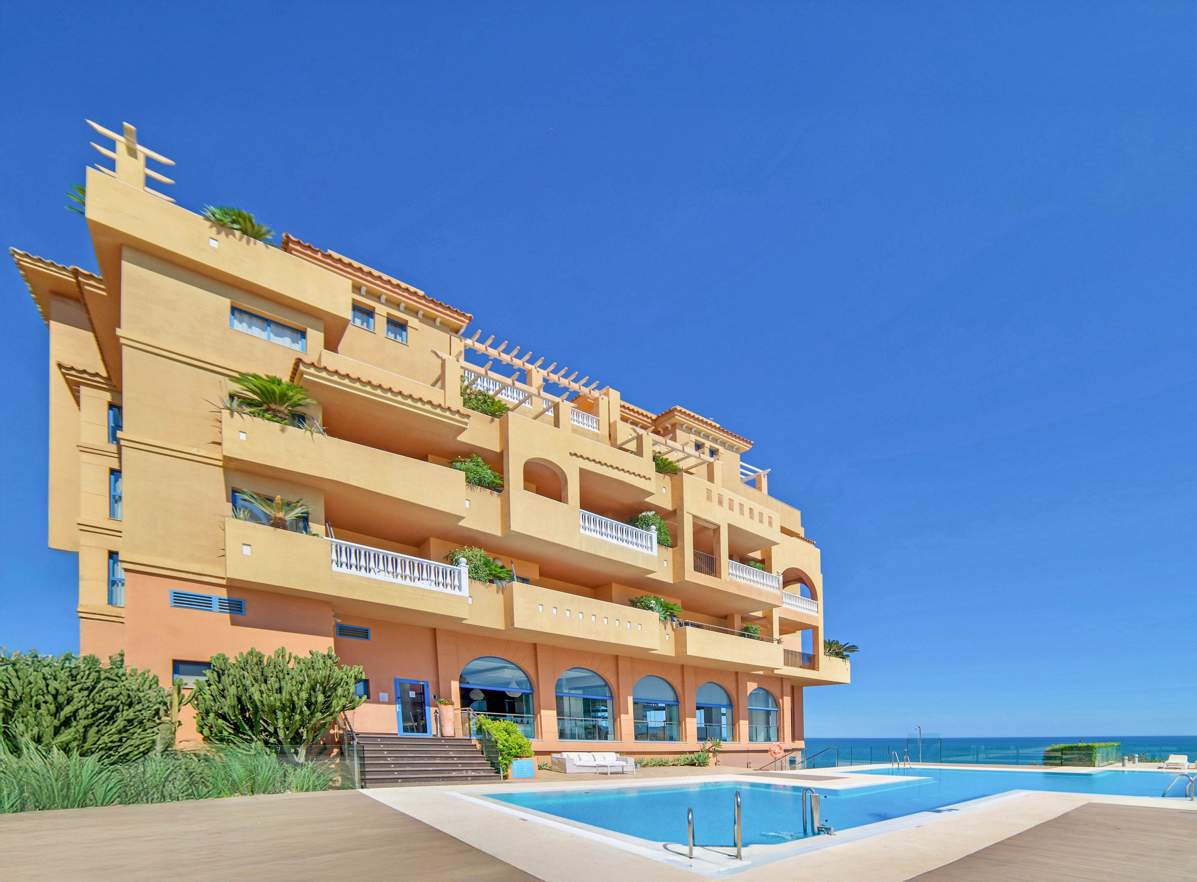 A multi-story apartment building stands prominently by the sea, featuring spacious balconies and Mediterranean architectural elements. The foreground showcases a clear blue swimming pool surrounded by a wooden deck, with lush greenery accenting the scene. The sky is bright and cloudless, enhancing the tranquil, vacation-like atmosphere. No visible text or numbers are present in the image.