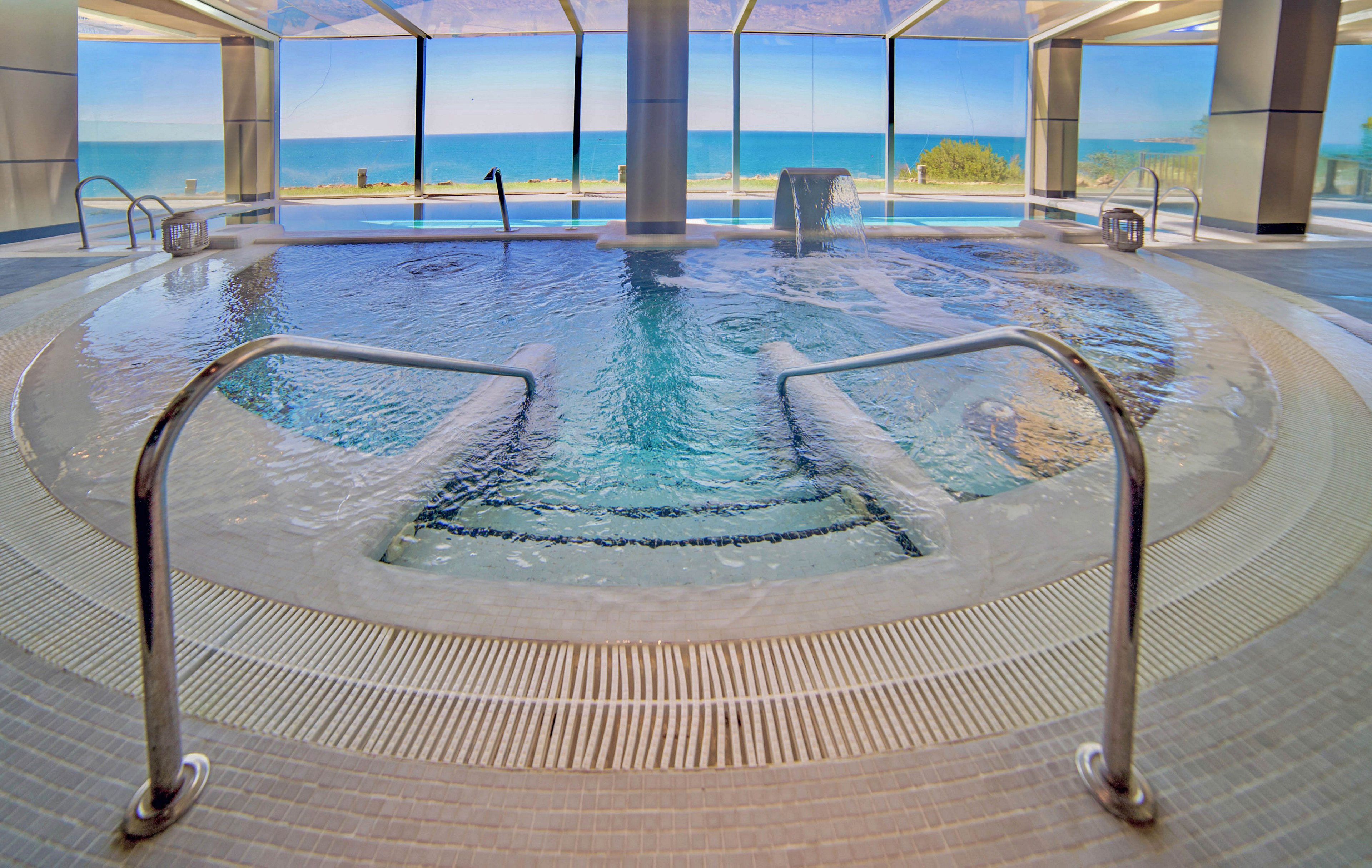 A circular indoor spa pool is featured in a modern, sunlit room with large floor-to-ceiling windows. The pool area offers a clear view of the ocean and sandy beach outside, creating a relaxing and luxurious atmosphere. Stainless steel handrails and tiled flooring add to the contemporary design. The setting is tranquil, with no people present, emphasizing calm and relaxation.