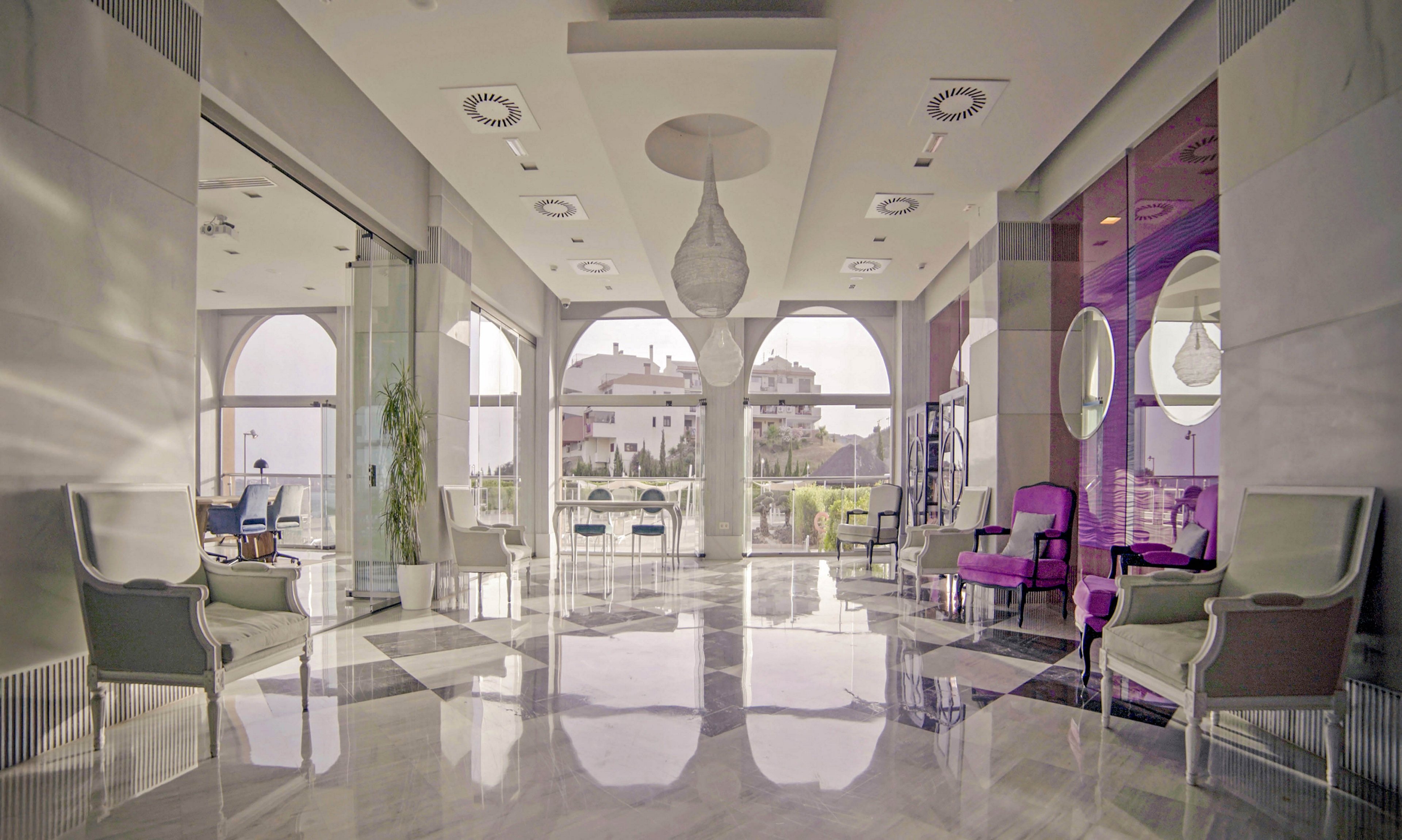 A spacious hotel lobby features modern armchairs and sofas arranged along polished marble floors. Large arched windows allow natural light to flood the space, highlighting the contemporary decor and subtle pastel color palette. The setting is tranquil and inviting, with a mix of neutral and purple tones adding sophistication. No people are present, emphasizing the serene atmosphere.
