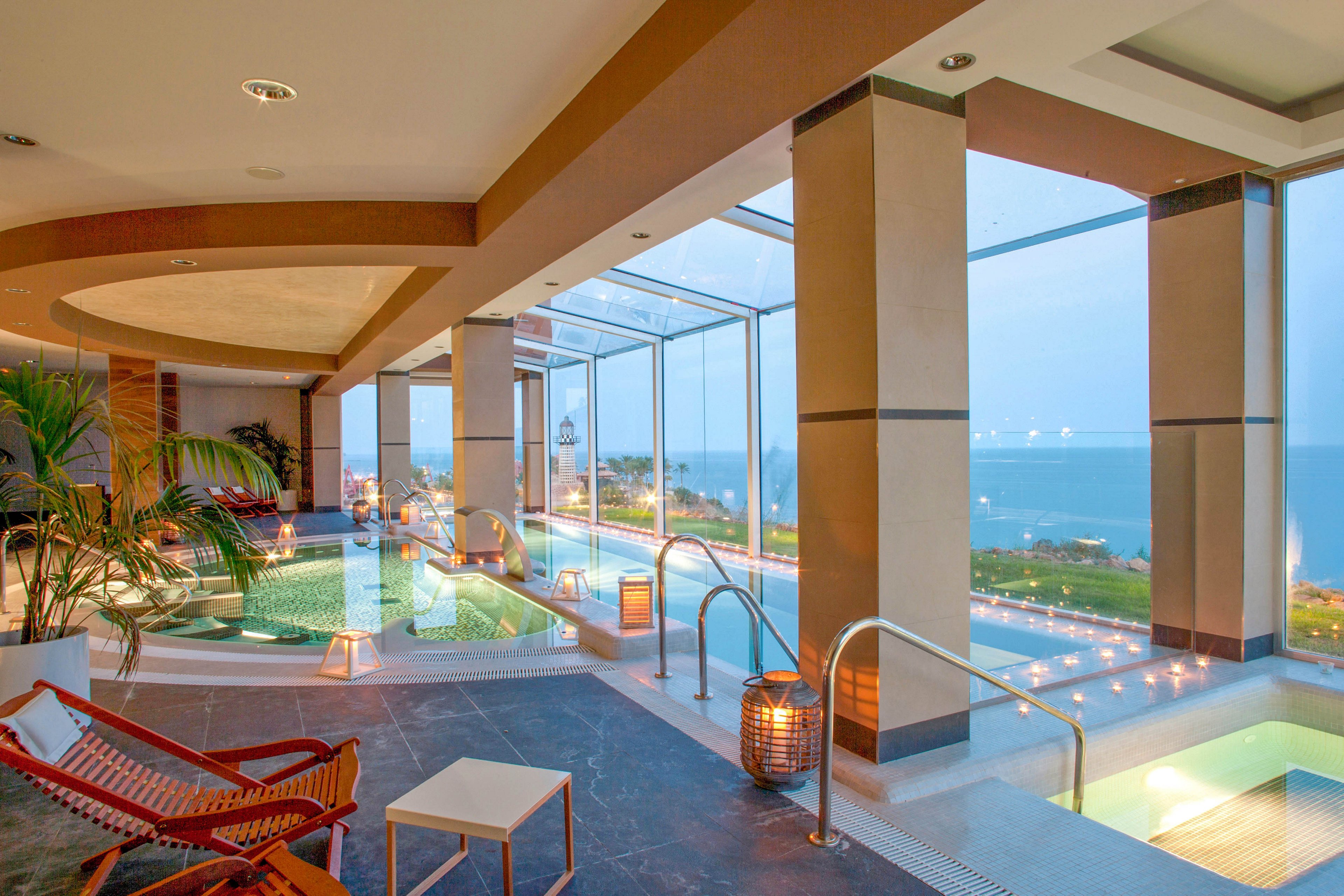 A modern indoor spa features multiple pools and relaxation areas with large glass windows offering a panoramic ocean view. The setting is tranquil, with soft lighting, candles, and comfortable lounge chairs enhancing the serene atmosphere. The interior design includes natural elements like plants and wooden accents, creating a calming retreat. No visible people or text are present in the image.
