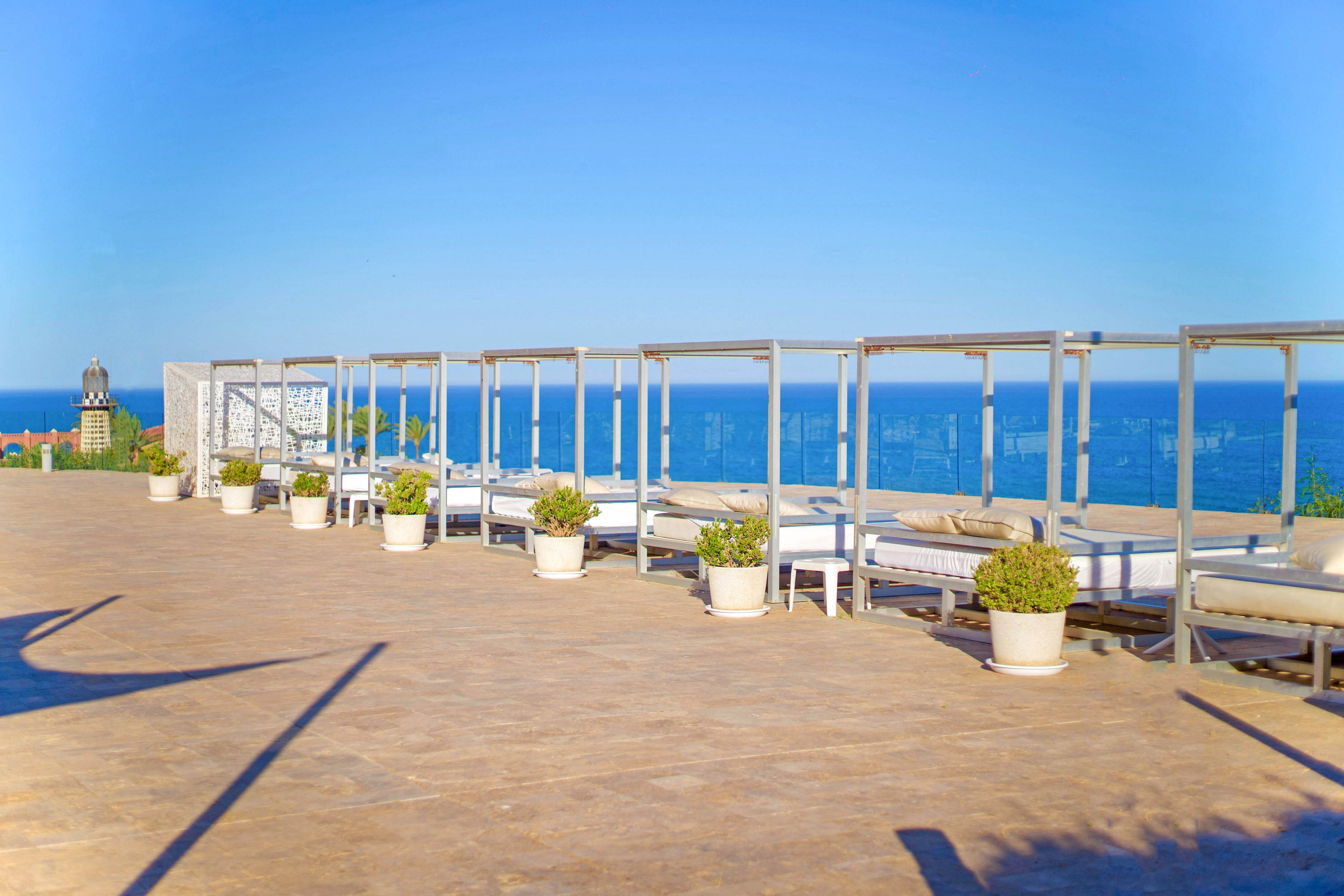 A spacious rooftop terrace features several canopy daybeds arranged in a row, each separated by potted plants. The setting overlooks a clear blue ocean under a bright sky, creating a serene and inviting atmosphere. The area is sunlit, with clean lines and a modern, minimalist style. No people are present, emphasizing tranquility and relaxation.