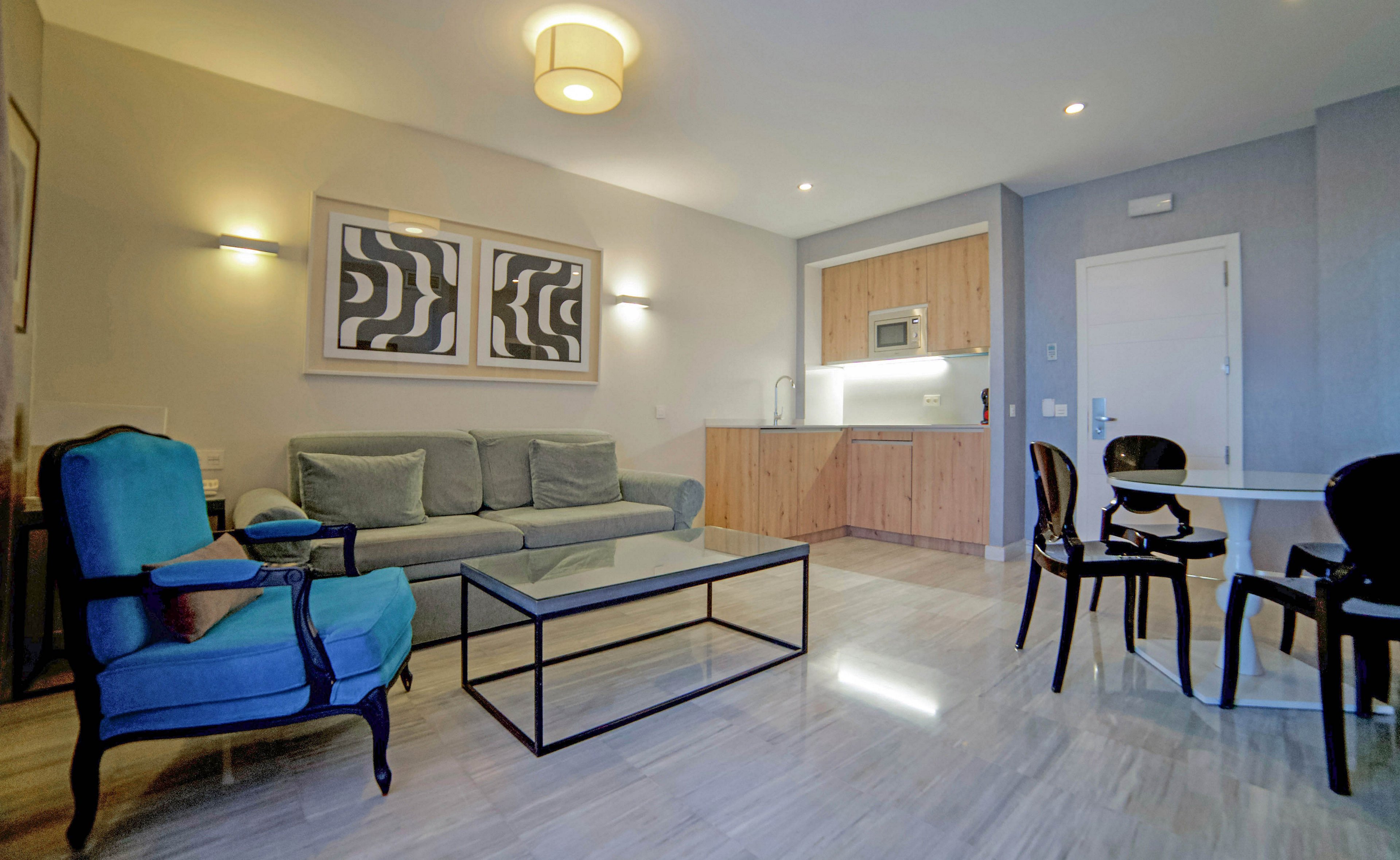 A contemporary living room features a gray sofa, glass coffee table, and blue armchair, set against a backdrop of abstract wall art. The open-plan space includes a compact kitchen with wood cabinetry and a small dining area with black chairs. The room is well-lit with ceiling and wall lights, and the overall palette is neutral with pops of blue and black.