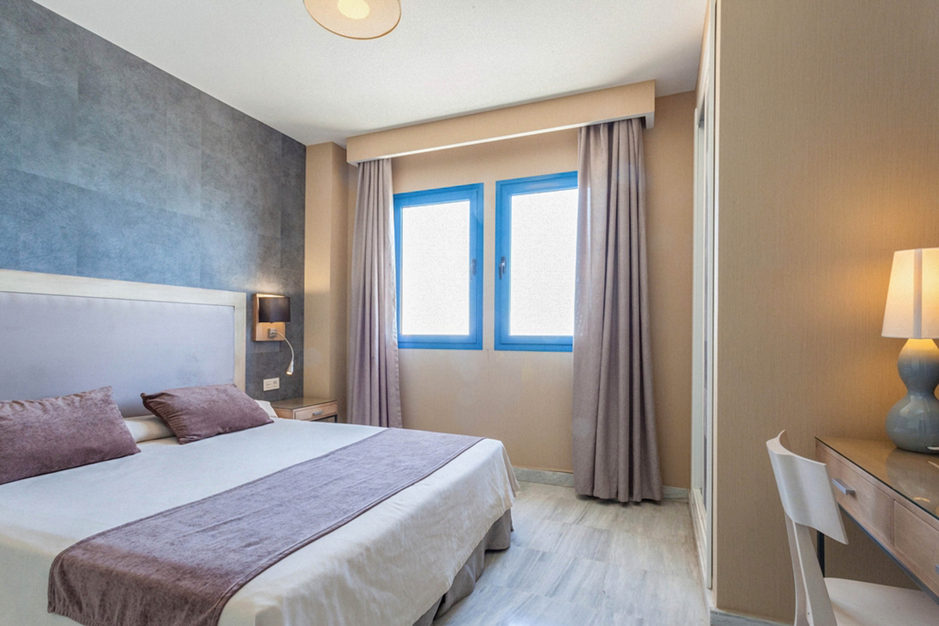 A contemporary hotel room features a double bed with mauve accents, two pillows, and a matching runner. The space is well-lit by natural light from two large windows with blue frames and taupe curtains. A desk with a lamp and chair sits to the right, while soft neutral tones and clean lines create a calm, inviting atmosphere. The room is unoccupied and appears tidy, emphasizing comfort and simplicity.