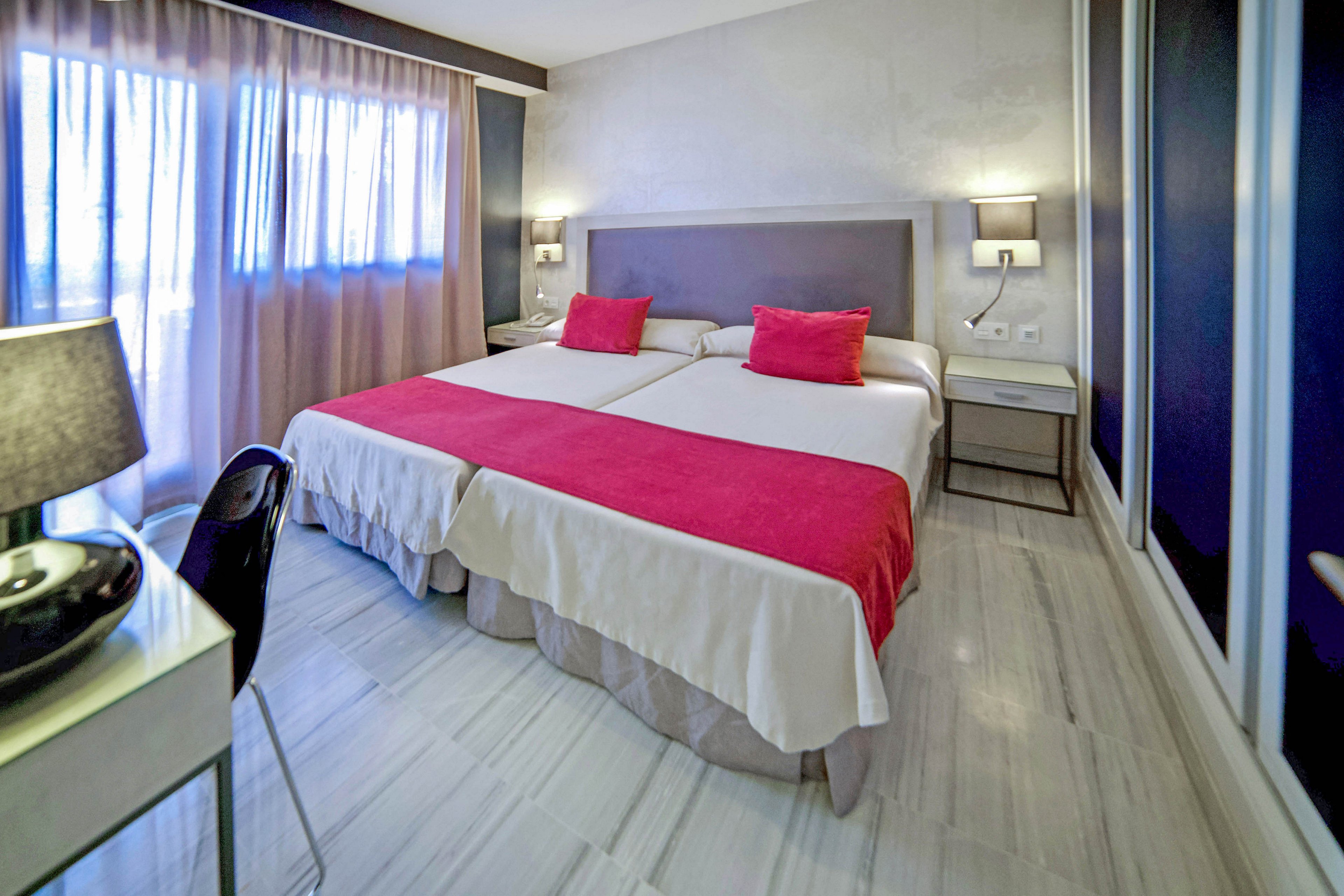 A contemporary hotel room features two twin beds pushed together, each topped with a bright pink runner and matching pillows. The space is well-lit with natural light streaming through sheer curtains, and the decor includes sleek furniture and neutral tones with pops of color. The room has a clean, minimalist style with a desk, chair, and bedside lamps. The overall mood is calm and inviting, ideal for relaxation or business travel.