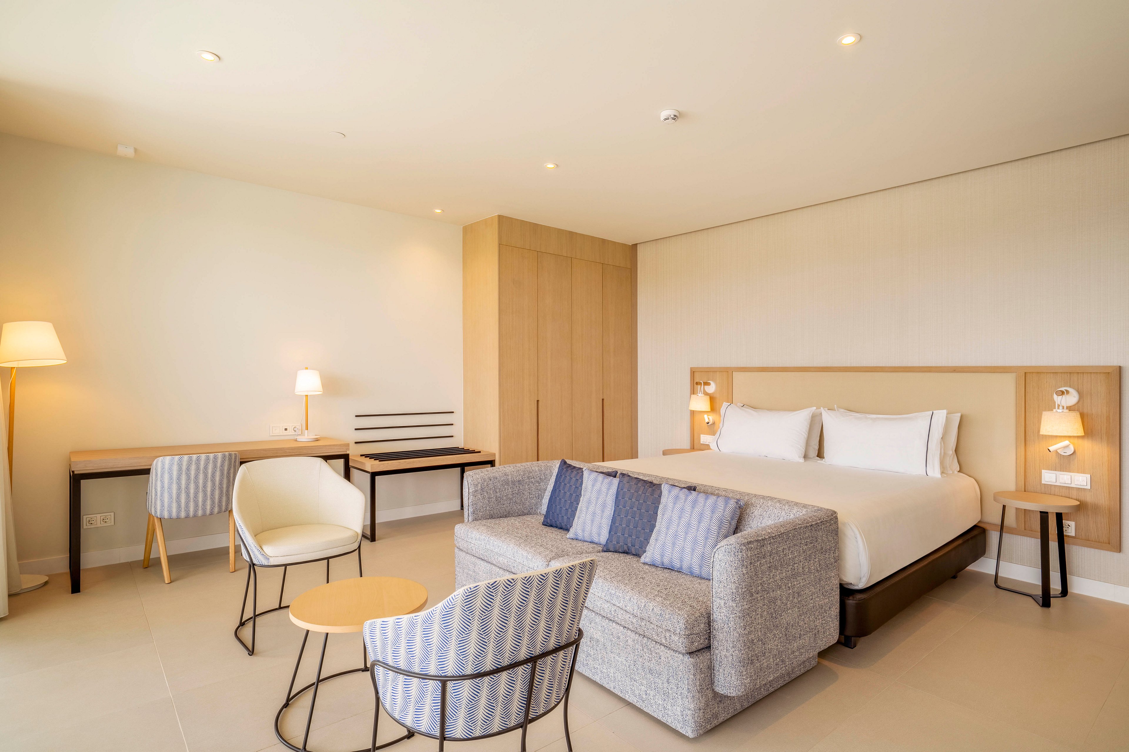 A spacious modern hotel suite features a large bed with crisp white linens and a light wood headboard. In front of the bed, a compact gray sofa with striped cushions faces a small round coffee table and matching striped armchairs. A wooden desk with a chair and floor lamp lines the wall, while a built-in wardrobe provides storage. Soft, warm lighting and neutral beige tones create a calm, minimalist atmosphere suitable for relaxation or work.