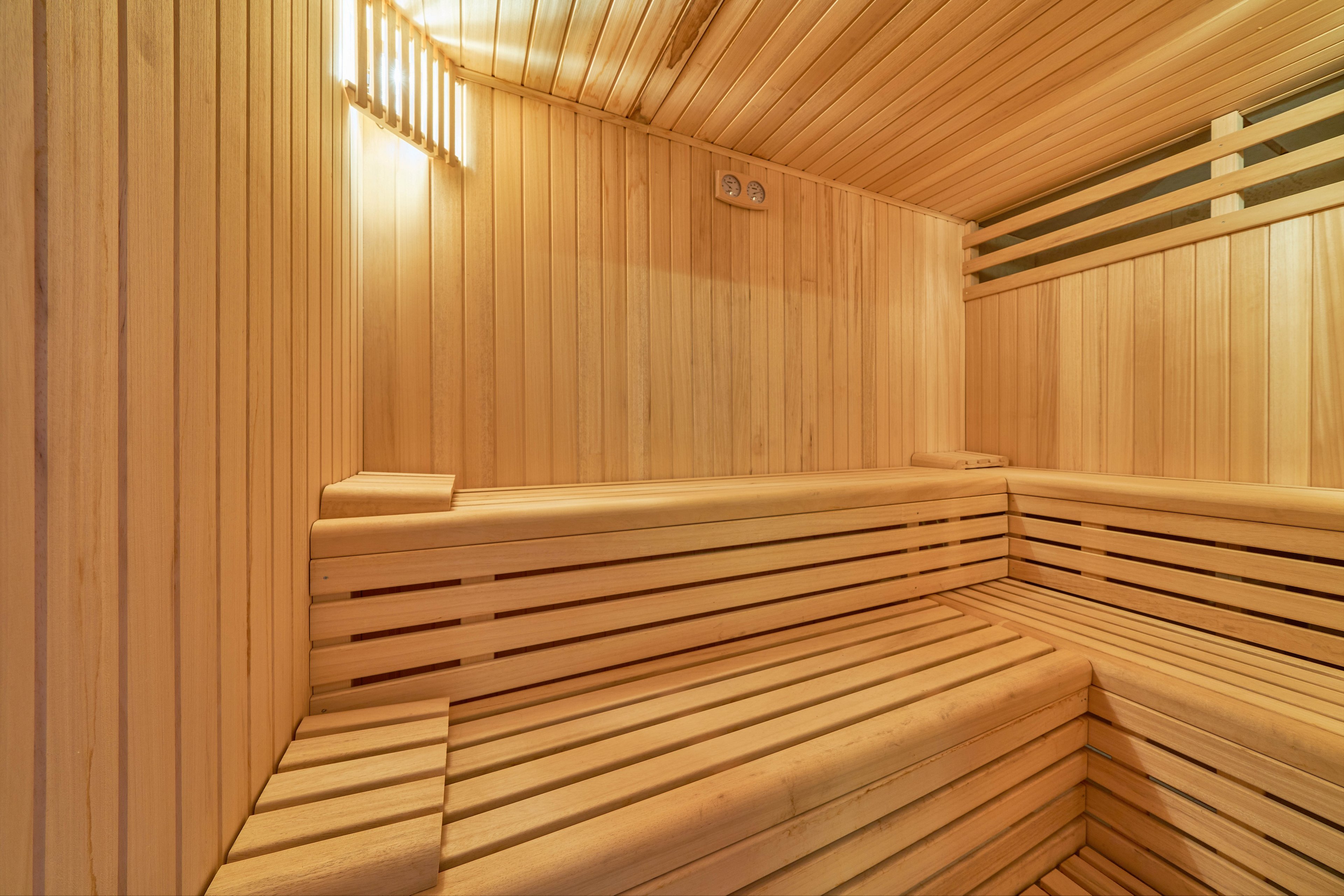 Warm-toned wooden sauna interior featuring tiered benches and slatted walls. The space is empty, highlighting the clean lines and craftsmanship of the wood paneling. Soft lighting creates a cozy, relaxing atmosphere suitable for spa or wellness settings. Ventilation slats and a wall-mounted control panel add functional details to the scene.