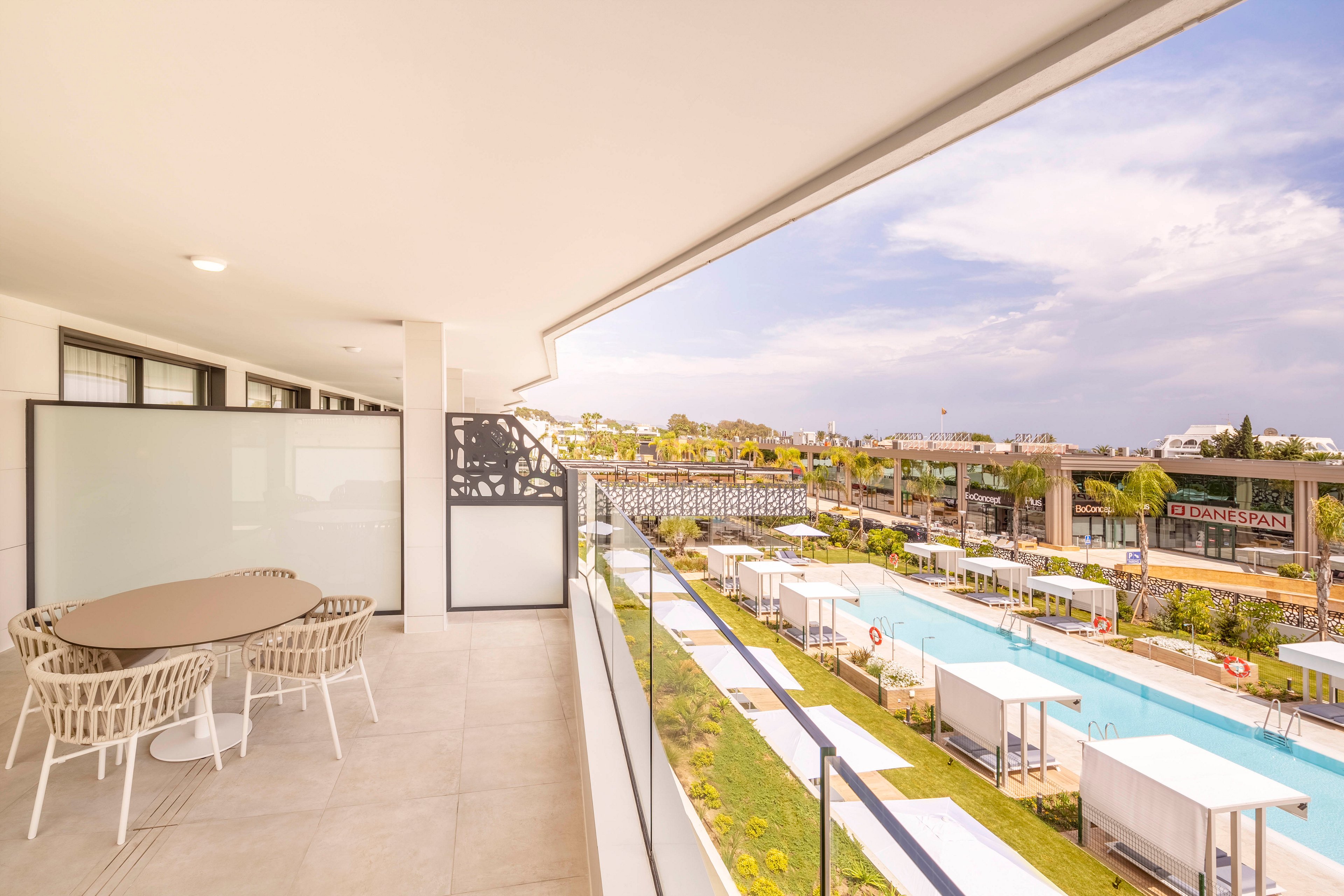 A spacious modern balcony with a round dining table and woven chairs overlooks a long rectangular swimming pool. White cabanas and sun loungers line the pool, creating a resort-like atmosphere. Contemporary low-rise buildings and palm trees frame the scene under a bright, partly cloudy sky. The overall palette is light and airy, with soft neutrals and turquoise water emphasizing a relaxed vacation mood.