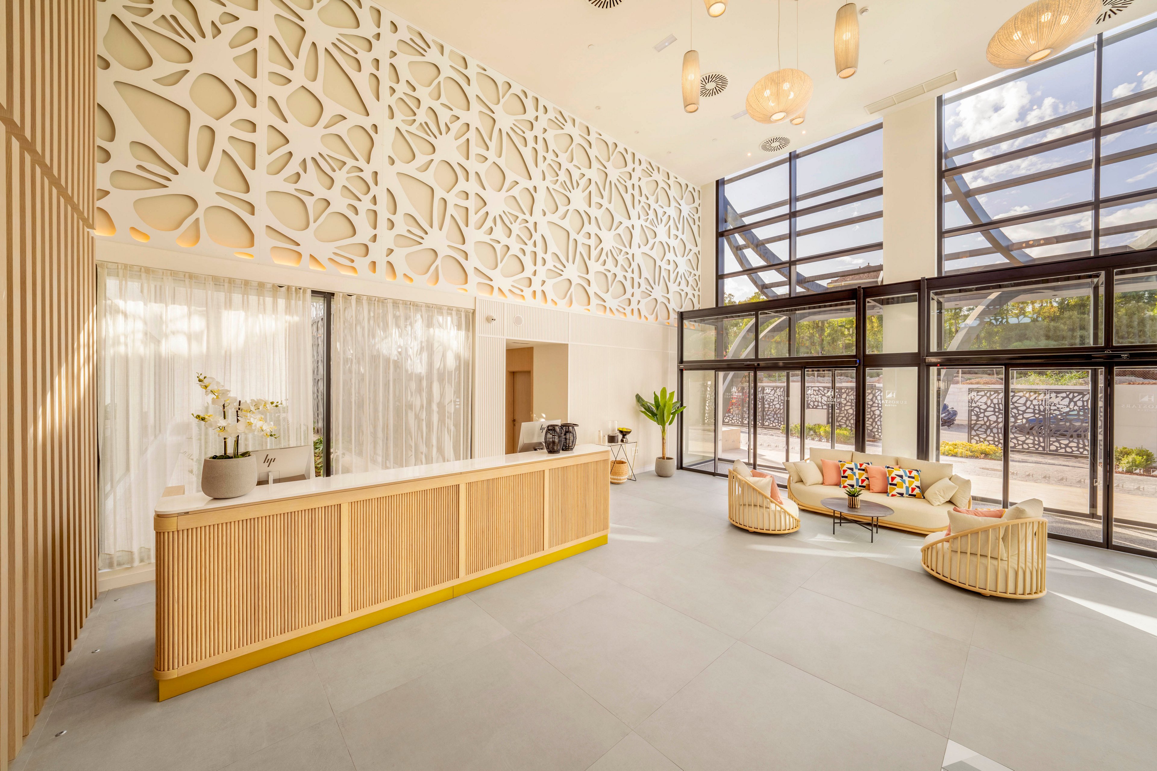 A spacious, contemporary lobby features a light wood reception desk with a potted orchid and decorative objects on top. The space is defined by a large patterned wall panel and floor-to-ceiling windows that let in abundant natural light. A small seating area with round chairs and a sofa sits near the glass entrance, accented by colorful cushions. Neutral tones, clean lines, and soft lighting create an airy, welcoming atmosphere.