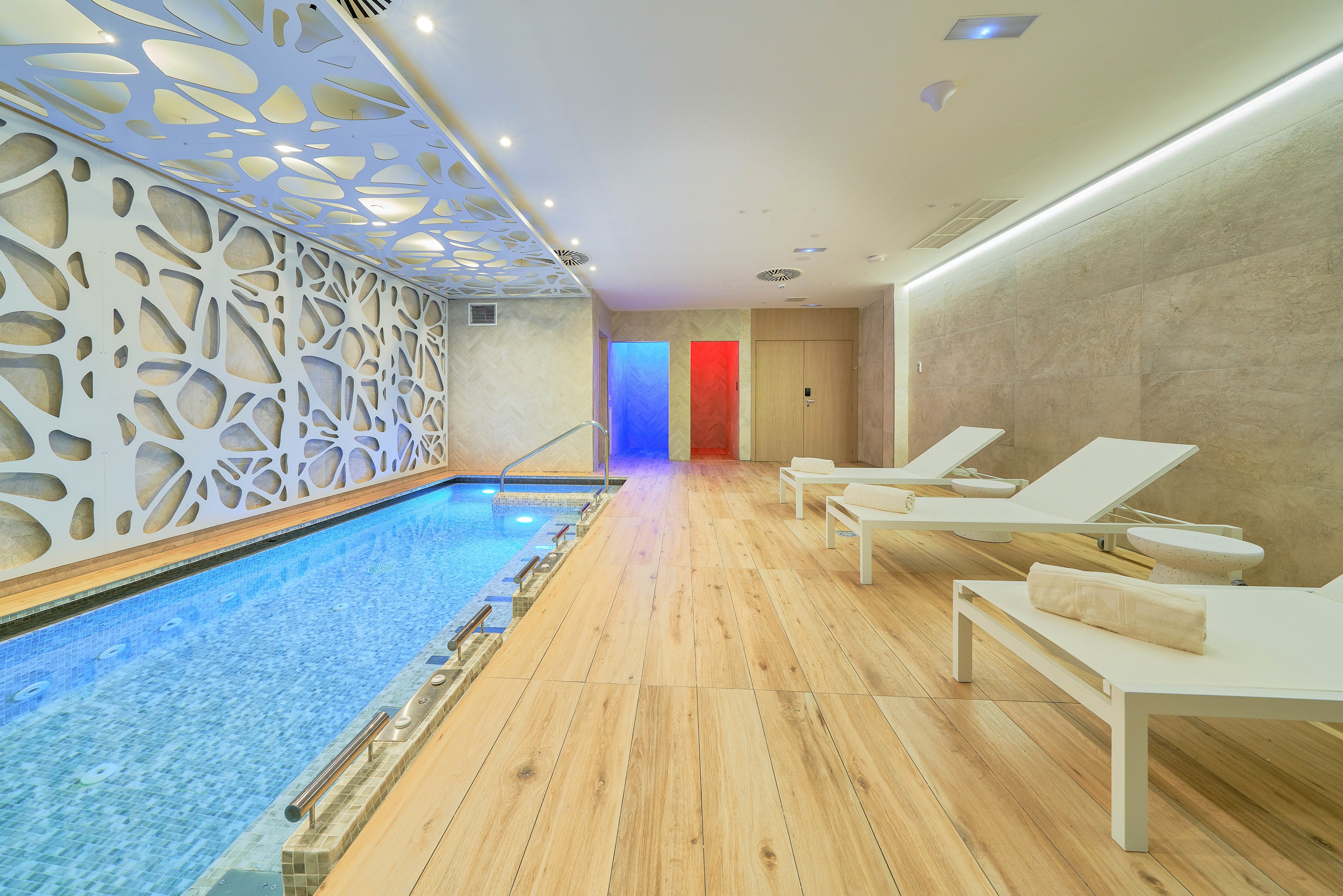 Bright contemporary spa interior featuring a narrow indoor pool with blue-tiled water. White lounge chairs with rolled towels line the wooden floor beside the pool. A decorative white wall with organic cutout patterns adds a stylish focal point. At the far end, illuminated panels in red, blue, and purple create a colorful, relaxing ambiance.