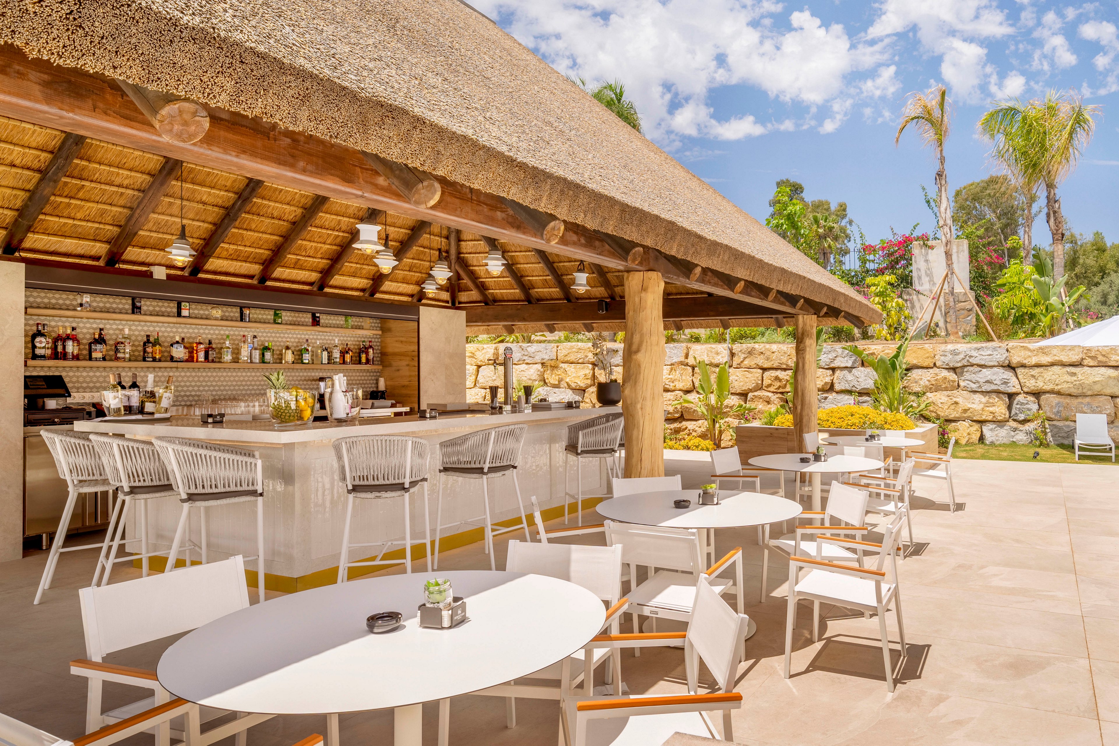 An open-air bar with a thatched roof features a long counter lined with modern barstools. White round tables and chairs are arranged on a spacious patio in front of the bar. Bottles and glassware are neatly displayed on shelves behind the counter, suggesting a well-stocked drink selection. Palm trees, stone walls, and a bright blue sky create a relaxed, tropical resort atmosphere.