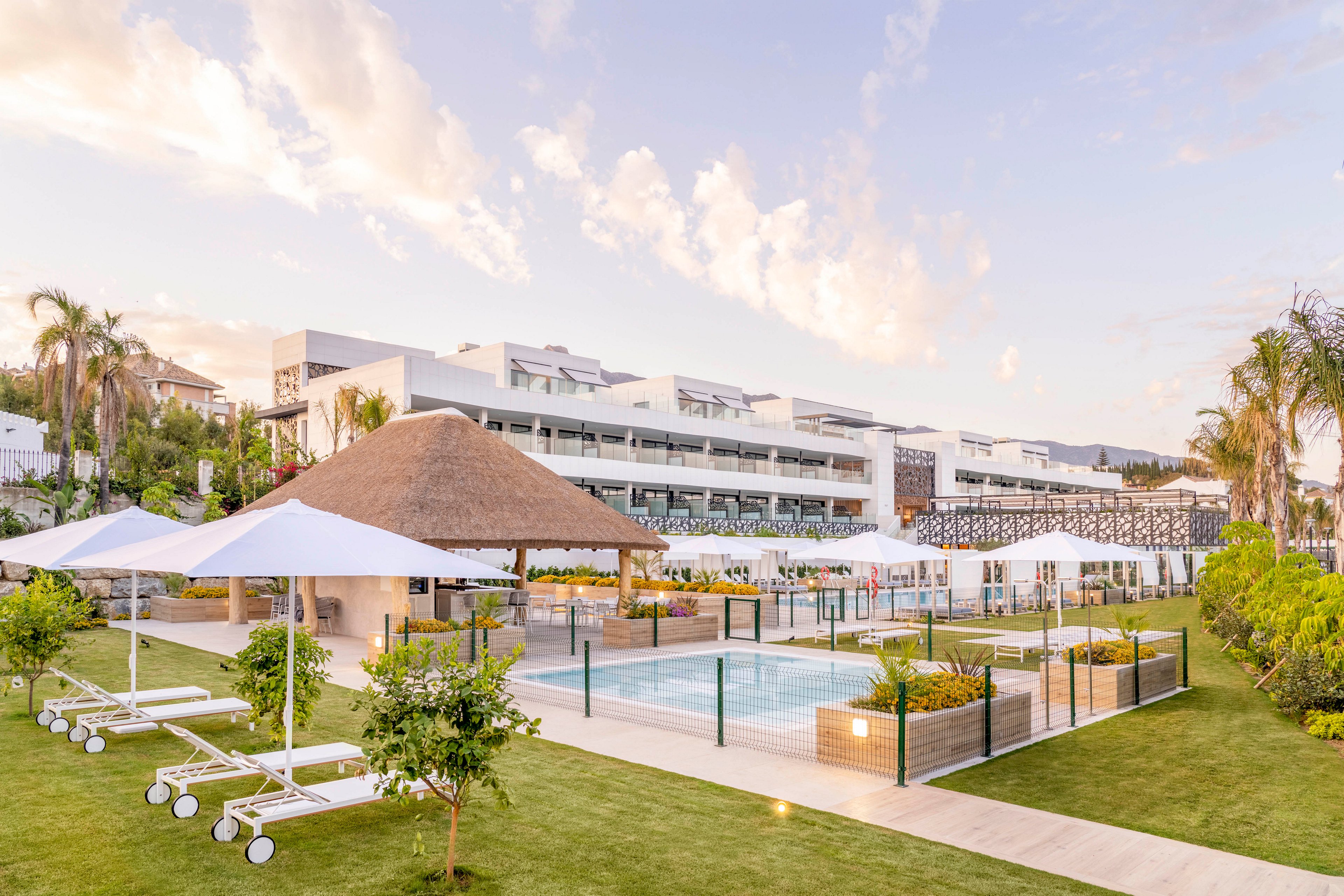 A contemporary resort complex features a landscaped poolside area with white sun loungers and umbrellas. A thatched-roof pavilion stands near the swimming pool, surrounded by manicured lawns and small citrus trees. The multi-story hotel buildings in the background have balconies and clean, minimalist architecture. Soft evening light and pastel skies create a calm, inviting atmosphere for relaxation.