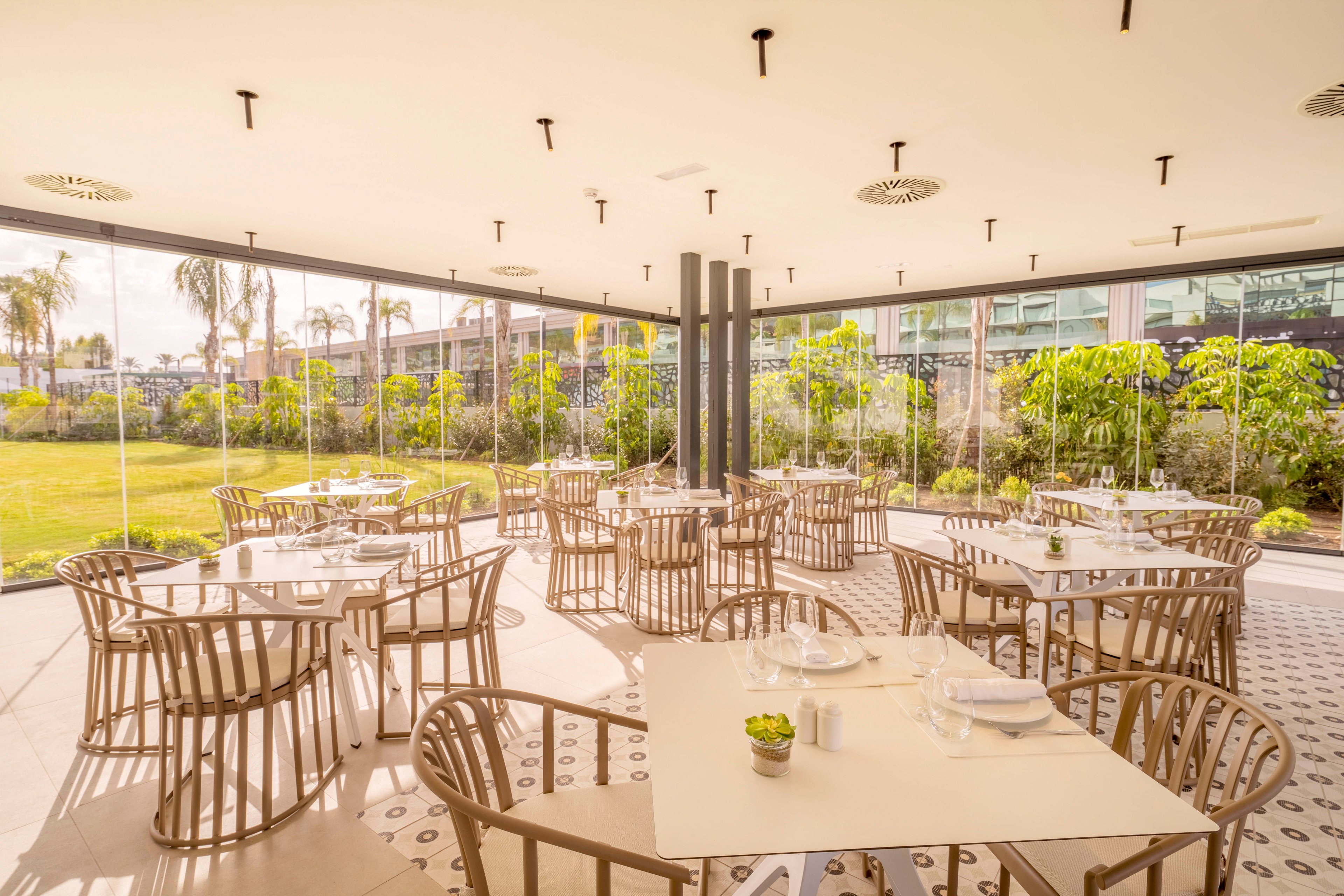 A contemporary restaurant terrace features neatly arranged tables and chairs in a bright, glass-enclosed space. The dining area overlooks a lush green garden with palm trees and manicured shrubs. Sunlight floods the room, highlighting the neutral tones of the furniture and patterned floor tiles. The setting feels airy and relaxed, ideal for casual dining or small gatherings.