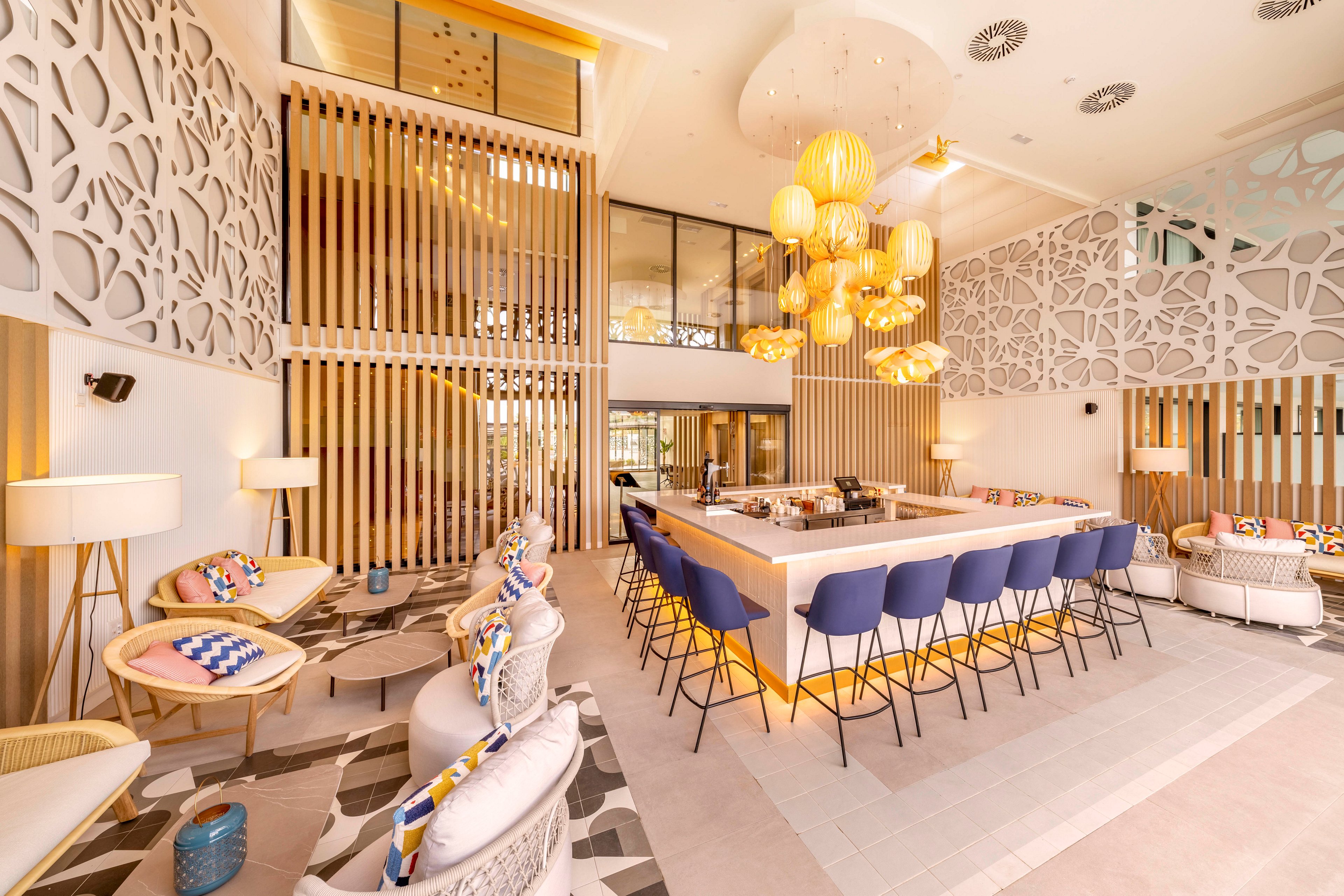 A contemporary hotel lounge features a central bar surrounded by blue barstools and cozy seating. The space is decorated with patterned wall panels, wooden slats, and soft neutral tones accented by colorful cushions. Warm pendant lights hang from the high ceiling, creating an inviting atmosphere. Large windows and glass partitions allow natural light to complement the ambient interior lighting.