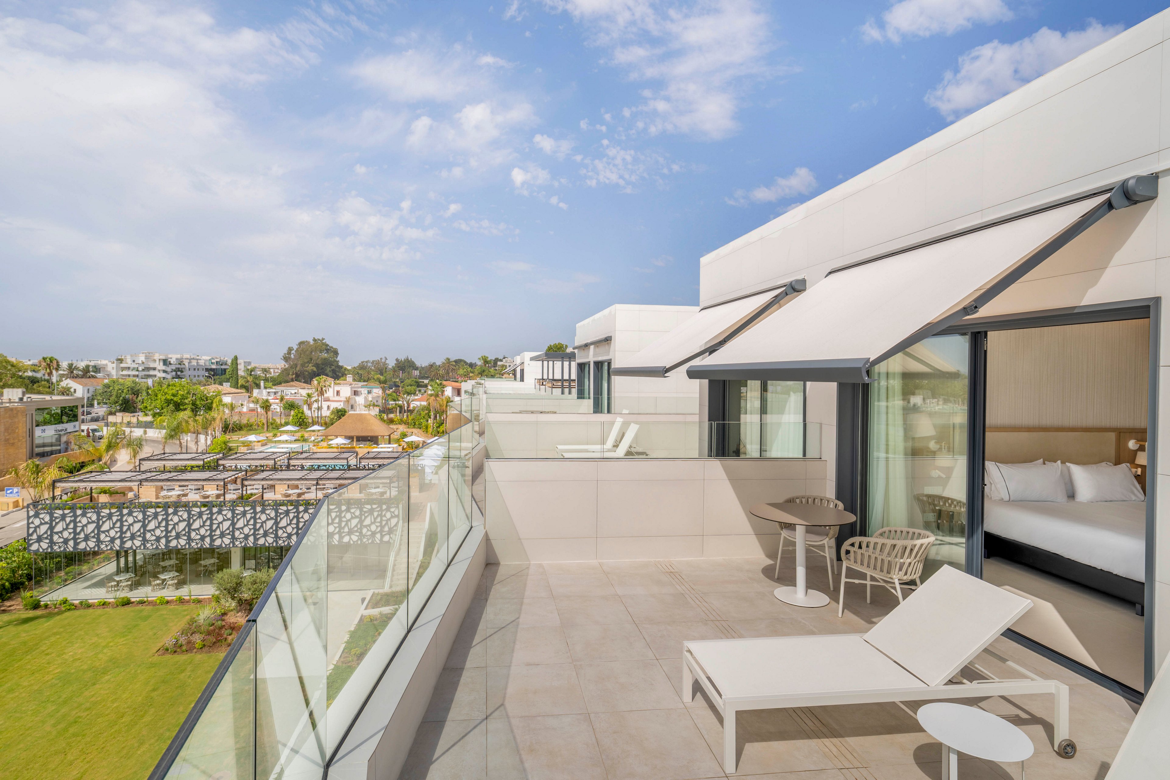A spacious modern hotel terrace features white lounge chairs and a small table overlooking a landscaped garden and cityscape. Large glass railings line the balcony, providing an open view of surrounding buildings and greenery. Sliding glass doors reveal a bright bedroom inside, while retractable awnings offer shade over the seating area. The scene is captured under a partly cloudy blue sky, creating a calm and inviting atmosphere.