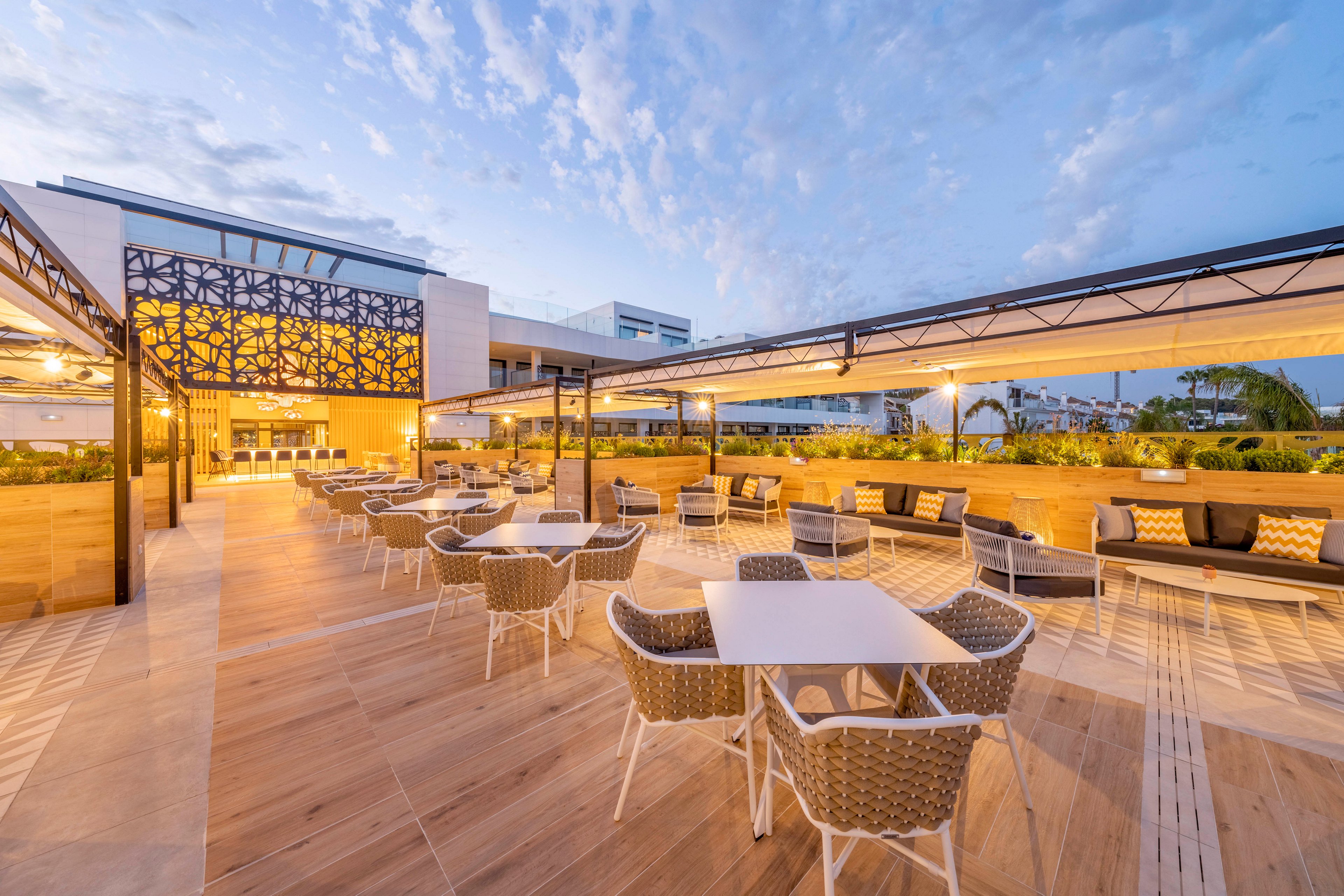 Spacious outdoor dining terrace with contemporary tables and woven chairs is shown under a soft evening sky. Warm architectural lighting highlights decorative lattice panels and wooden flooring. Long benches with cushions line the perimeter, creating a relaxed lounge area. The setting appears calm and inviting, ideal for al fresco dining or social gatherings.