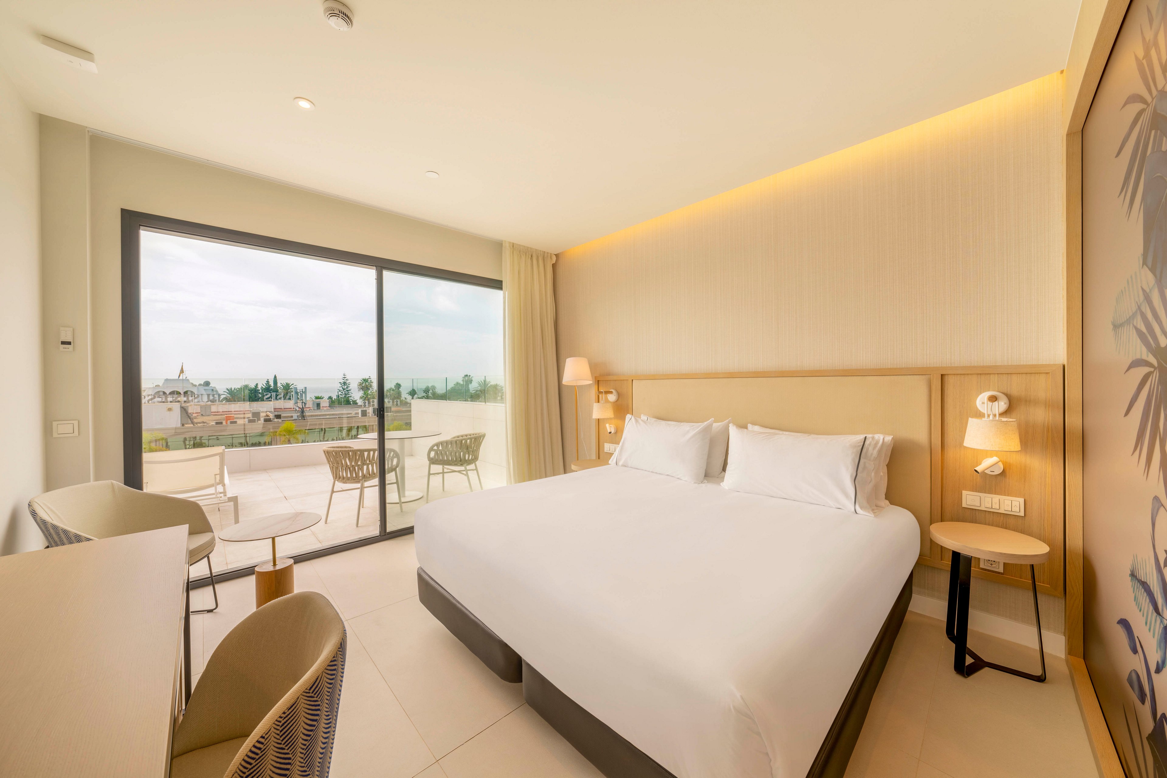 A contemporary hotel room features a large neatly made bed with white linens and soft lighting. Floor-to-ceiling glass doors open onto a balcony with outdoor chairs and a view of surrounding buildings and greenery. Neutral tones, minimalist furniture, and clean lines create a calm, inviting atmosphere. The space appears well-lit with natural daylight and subtle warm accent lighting along the ceiling.