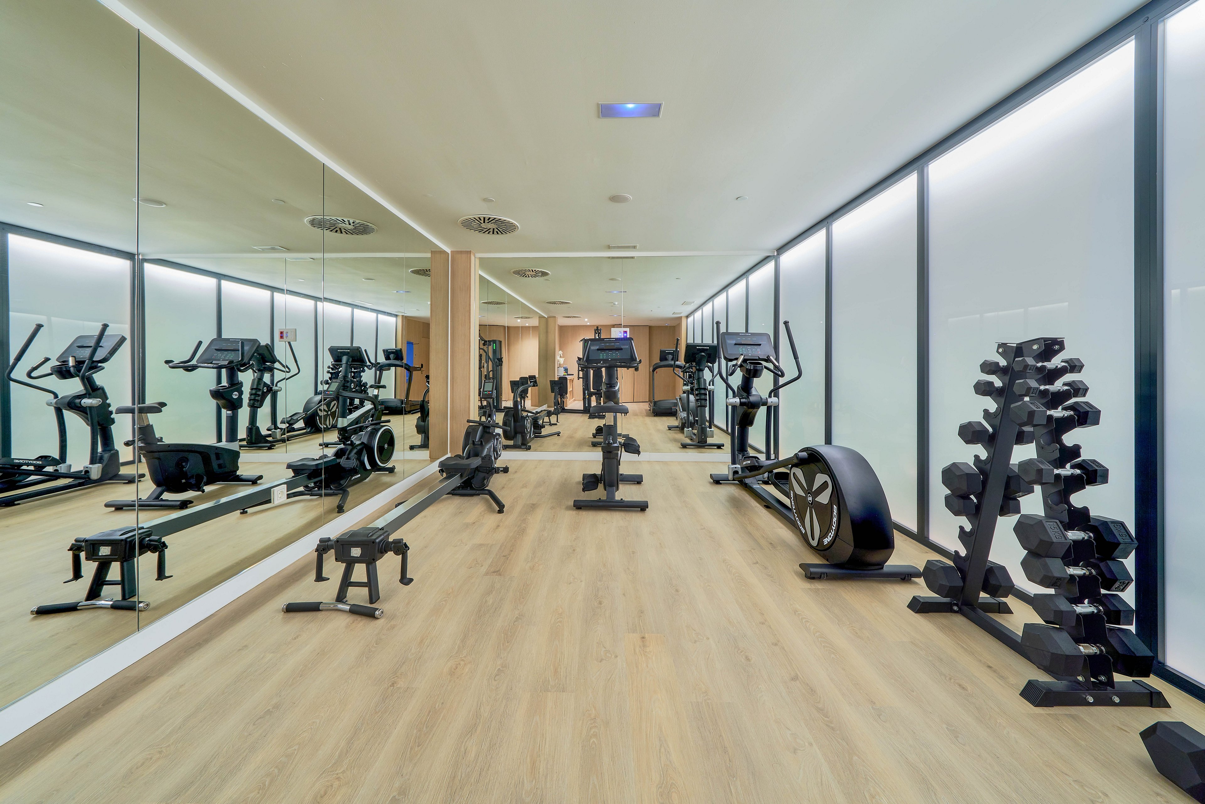 A bright, modern gym interior features various cardio and strength training equipment neatly arranged along a wooden floor. Treadmills, exercise bikes, and rowing machines line the mirrored wall, reflecting the spacious workout area. On the right side, a rack of dumbbells and other free weights is visible near frosted glass partitions. The clean, minimalist design and ample lighting create an inviting space for indoor exercise and fitness training.
