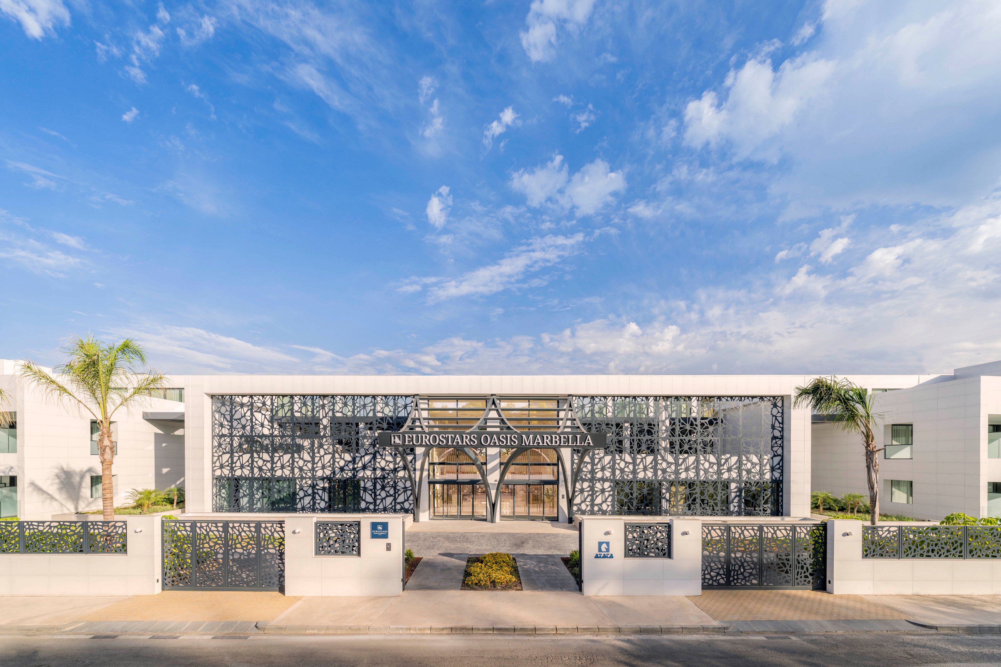 Contemporary hotel building with a symmetrical facade is shown under a bright blue sky. The entrance features decorative metal lattice panels and large glass windows. Palm trees and low hedges frame the front walkway and perimeter wall. The text 'EUROSTARS OASIS MARBELLA' is clearly visible above the main entrance, indicating the hotel name.