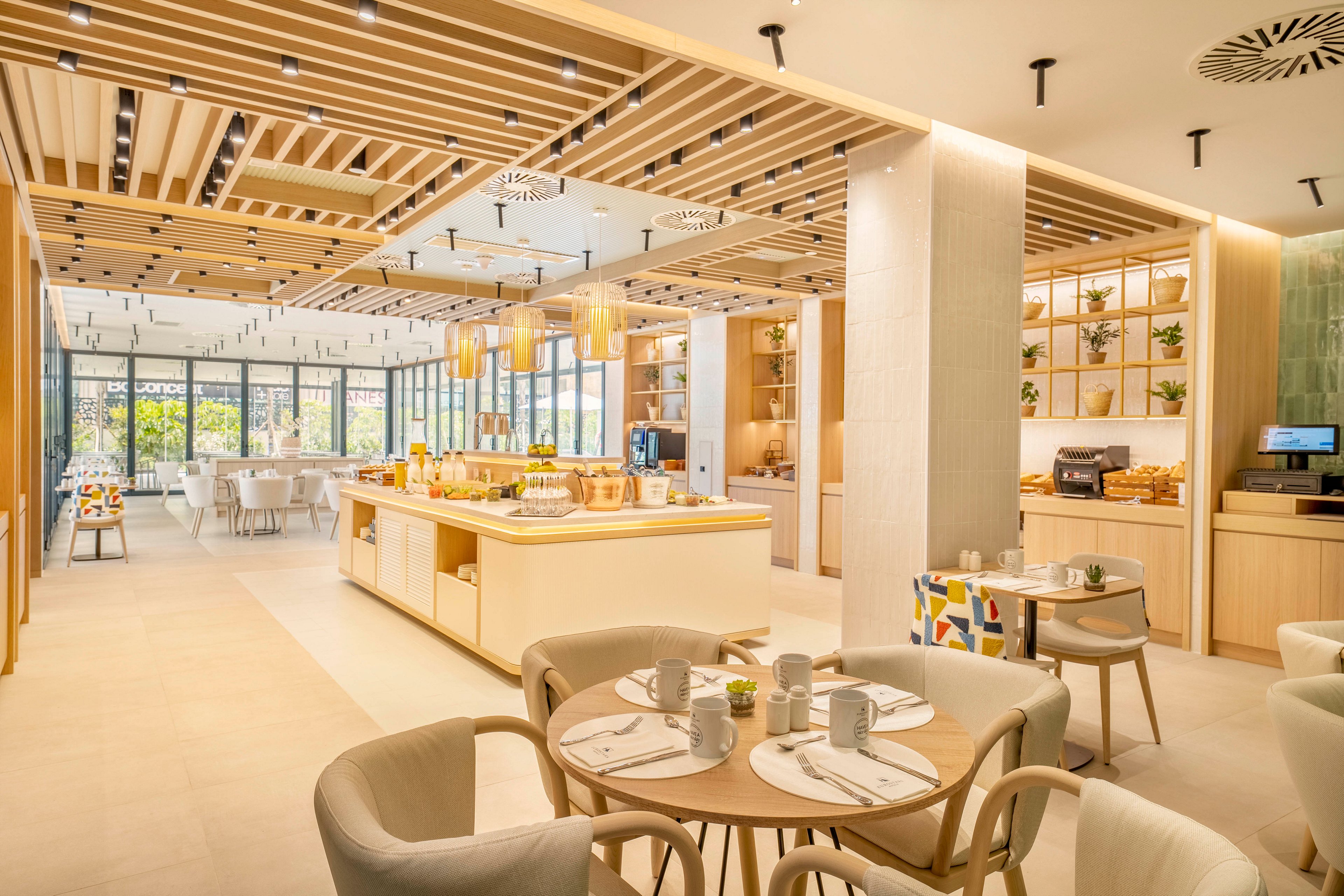 A spacious modern restaurant interior features light wood furnishings and neutral tones. Round tables are set with plates, cups, and cutlery in the foreground, while a central buffet island displays food and drinks. Large floor-to-ceiling windows bring in natural light and offer views of greenery outside. Ceiling slats with recessed lighting and decorative shelving with plants create a warm, inviting atmosphere.