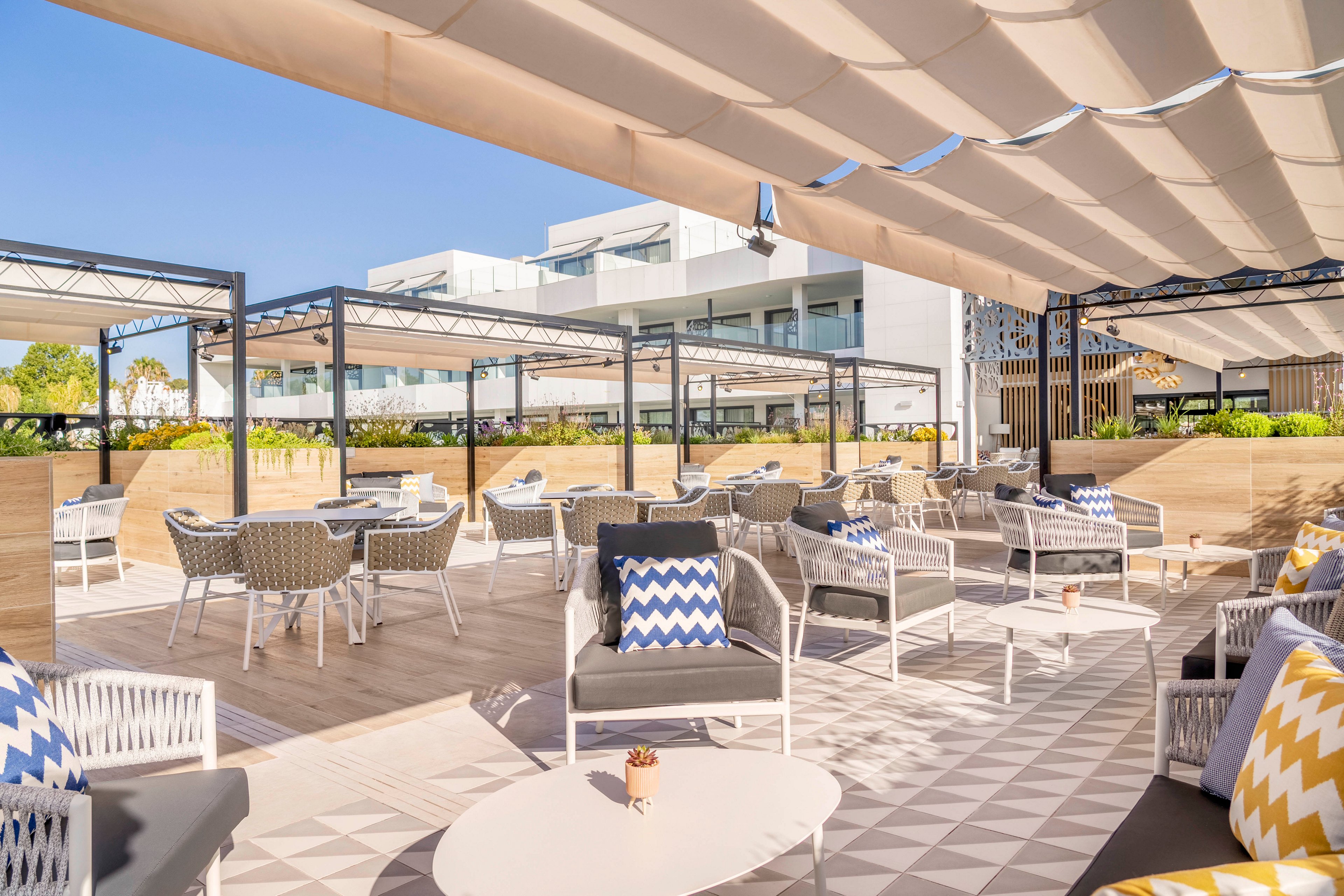 Contemporary outdoor seating area features woven chairs and cushioned sofas arranged around low tables. A large pergola-style sunshade structure covers the terrace, providing shade under clear blue skies. Geometric chevron-pattern pillows in blue, yellow, and white add color to the neutral flooring and furniture. In the background, modern white buildings and greenery frame the relaxed, resort-like setting.