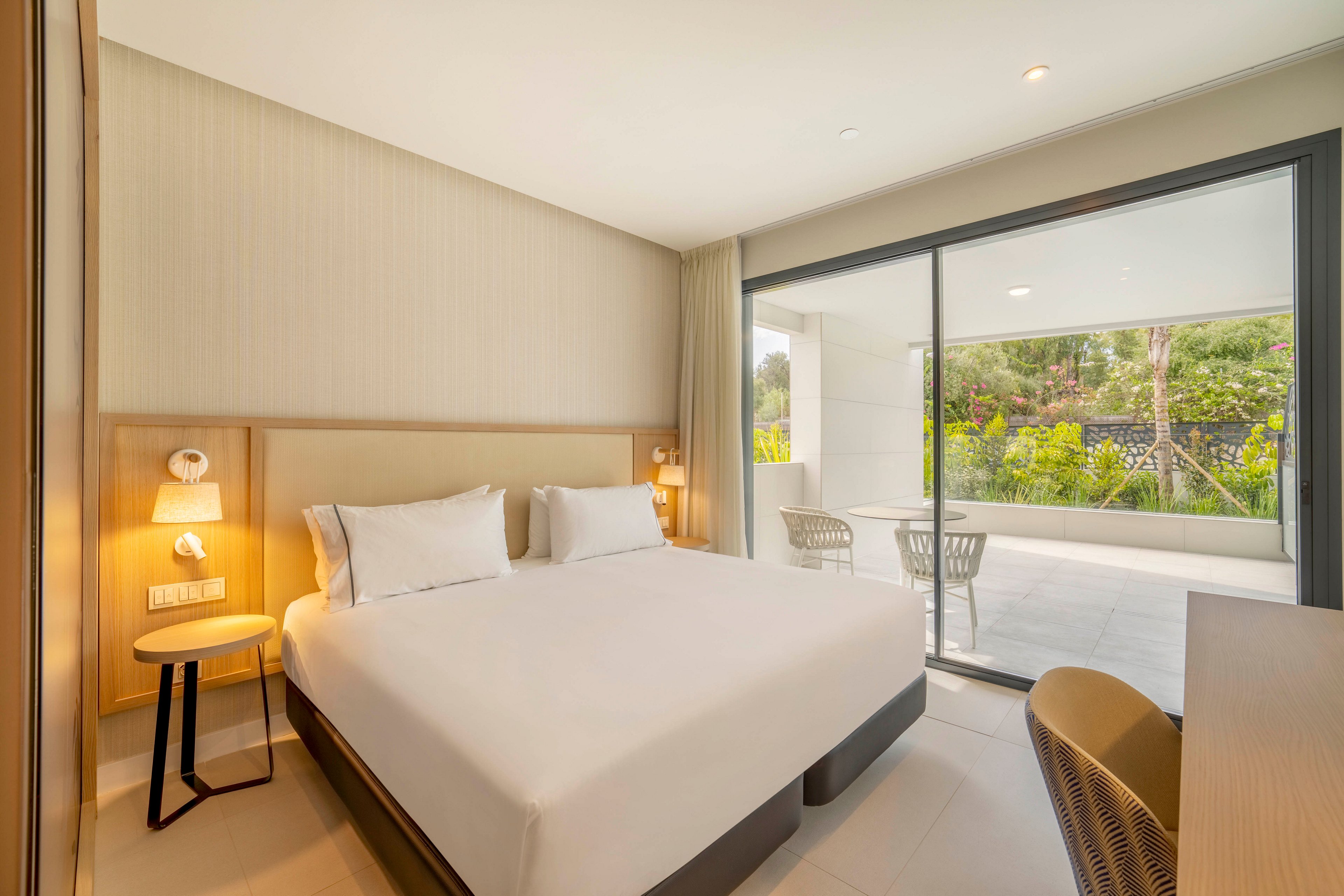 A bright modern hotel bedroom features a large bed with crisp white linens and minimalist wooden furniture. Warm bedside lamps illuminate the neutral-toned walls and headboard. A sliding glass door opens to a spacious terrace with outdoor chairs and a table. Beyond the terrace, greenery and a stone wall create a calm, private atmosphere.
