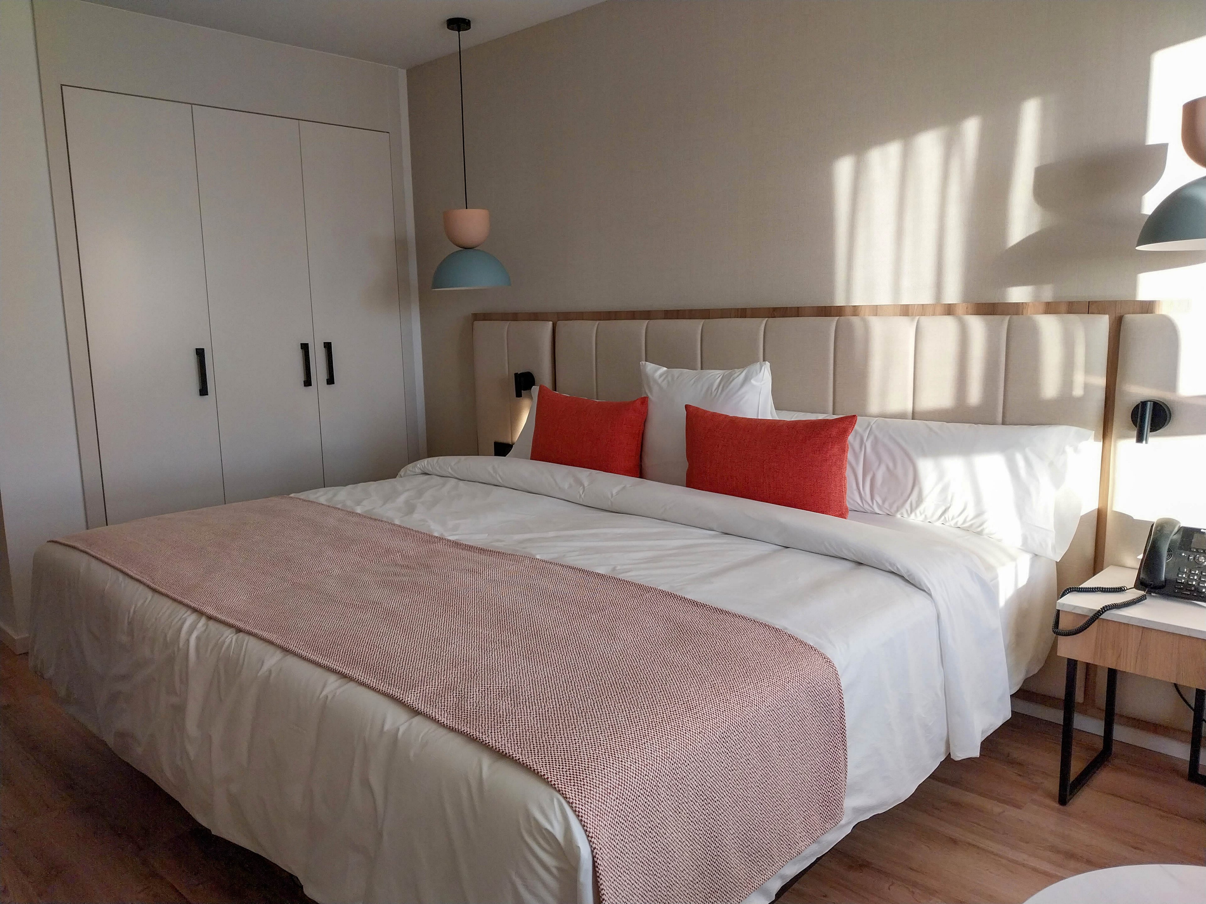 A contemporary bedroom features a large double bed with white linens and a soft pink throw blanket. Bright orange accent pillows add color against a neutral upholstered headboard. Built-in white wardrobes with black handles line the wall, and minimalist pendant lights hang above the bedside tables. Sunlight streams through an unseen window, creating a warm and inviting atmosphere with clean, modern decor.