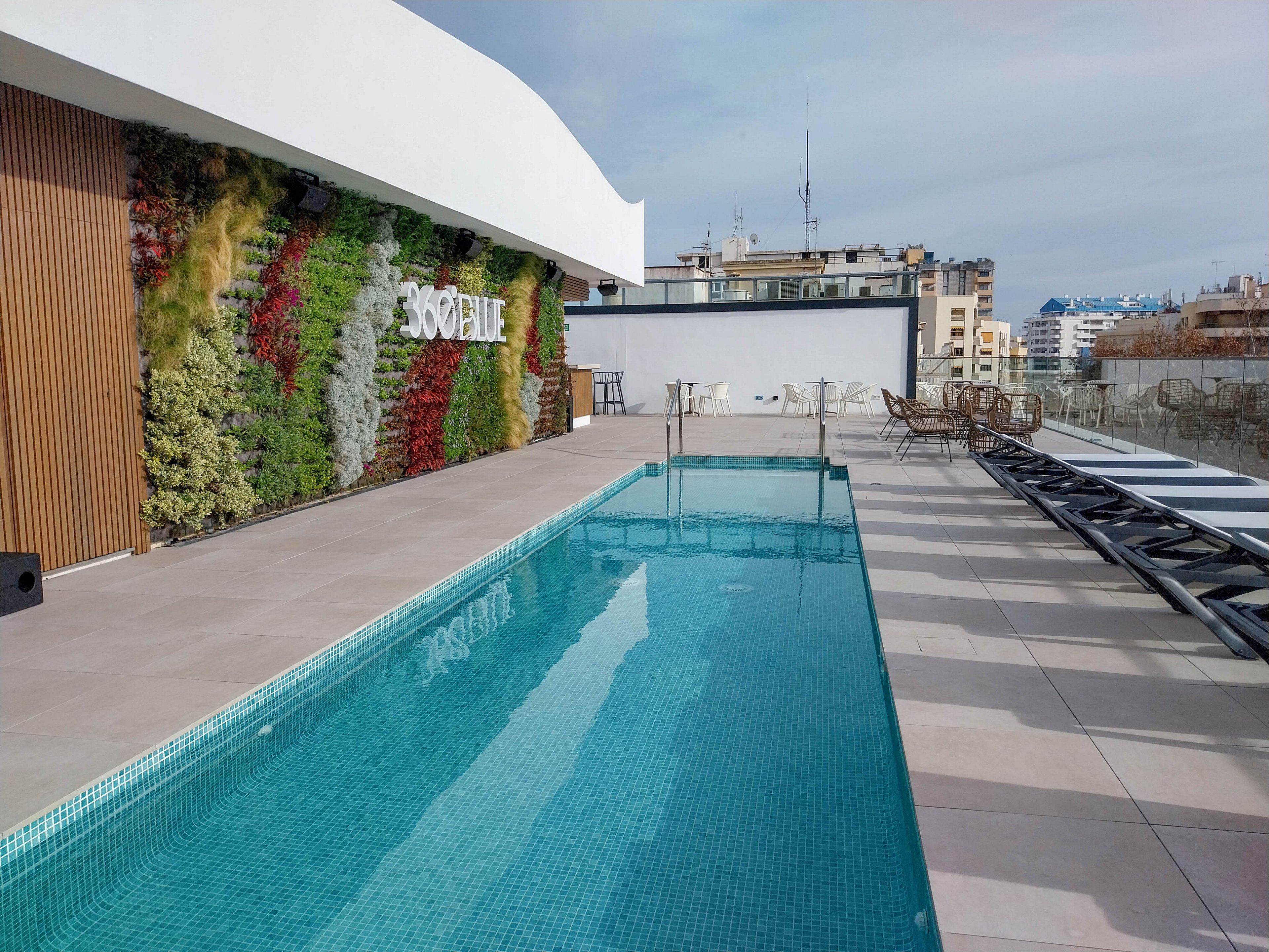 A sleek rooftop swimming pool stretches alongside a tiled terrace in an urban setting. One side features a tall vertical garden wall with colorful plants and the word "SOCIETE" clearly visible. Sun loungers line the opposite side of the pool, and outdoor chairs and tables sit near the far end. City buildings and antennas are visible in the background under a bright, partly cloudy sky.