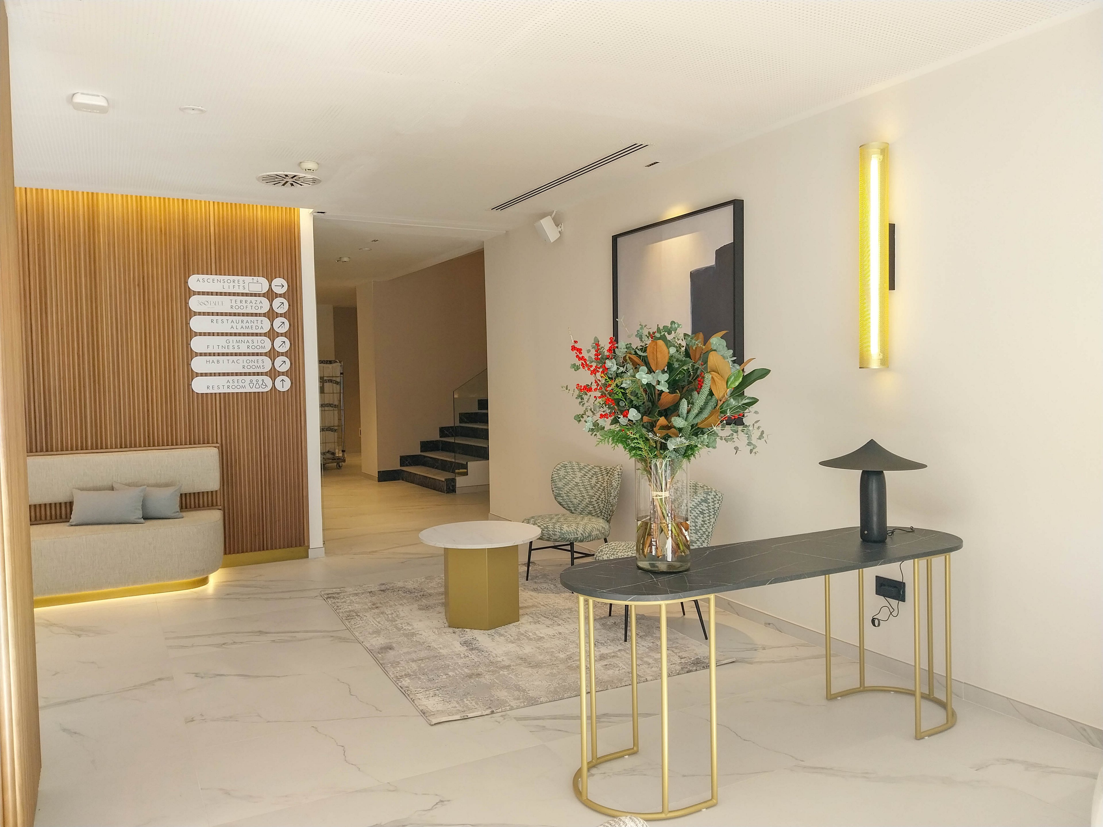 A contemporary lobby features a sleek console table with a glass vase of colorful flowers as the focal point. The space includes a light-toned sofa nook against vertical wooden slats and a round coffee table on a soft rug. Minimalist wall art, a staircase in the background, and warm wall-mounted lighting create a refined, welcoming atmosphere. The overall design emphasizes clean lines, neutral tones, and subtle gold accents.