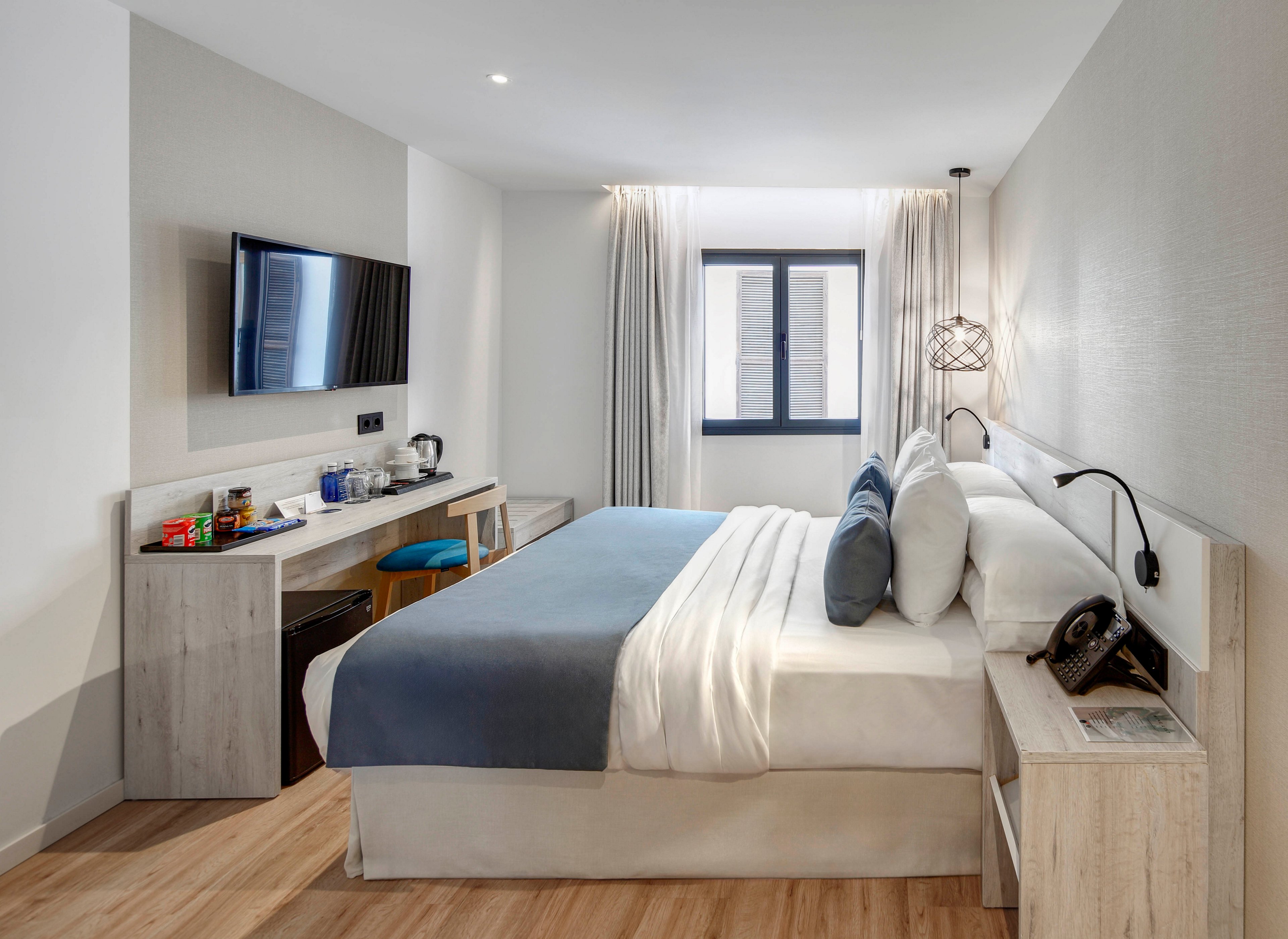 A contemporary hotel room features a neatly made double bed with white linens and a blue throw blanket. The space includes a wall-mounted flat-screen television opposite the bed and a light wood desk with a minibar and coffee station. Natural light enters through a central window framed by sheer curtains, brightening the neutral-toned walls and wooden floor. Decorative pillows, a bedside telephone, and a modern pendant lamp add comfort and style to the minimalist interior.