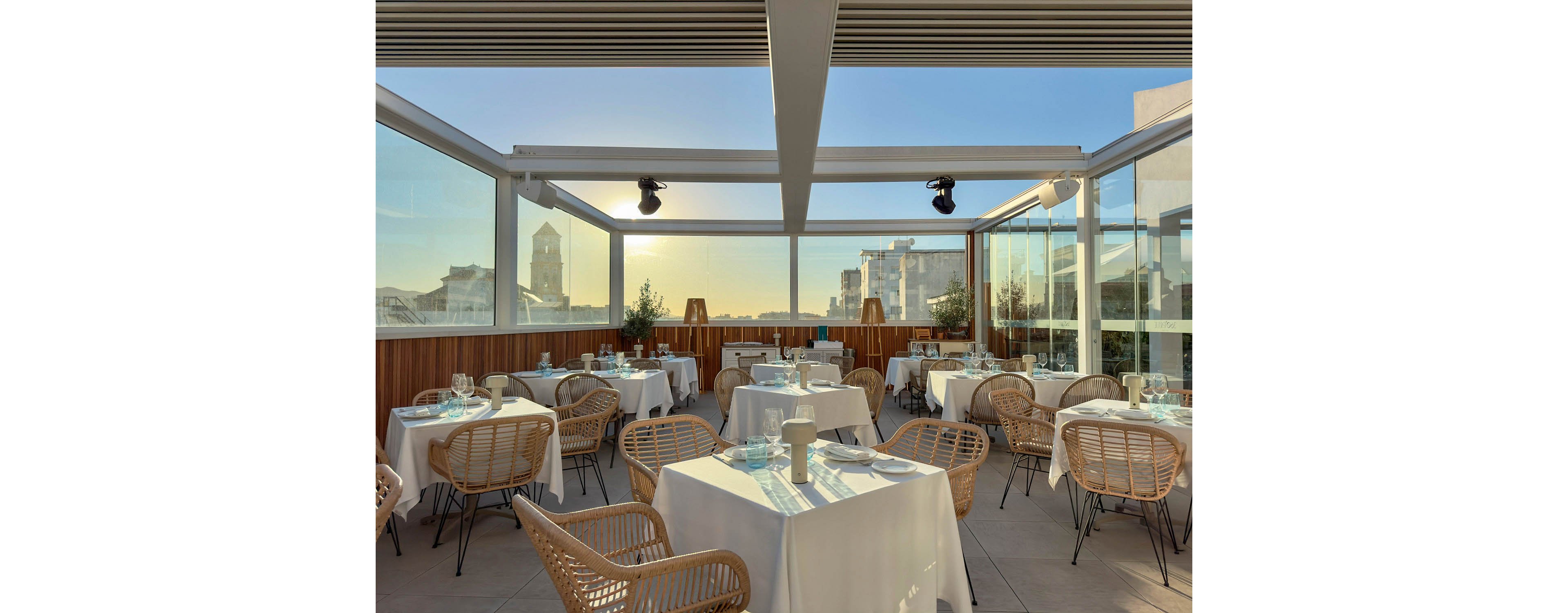An elegant rooftop restaurant is set with white tablecloths and wicker chairs under a modern pergola structure. Glass walls surround the terrace, offering expansive views of nearby city buildings and a distant tower. Tables are neatly arranged with glassware and plates, ready for guests in the warm evening light. The overall atmosphere feels relaxed, contemporary, and inviting for outdoor dining.