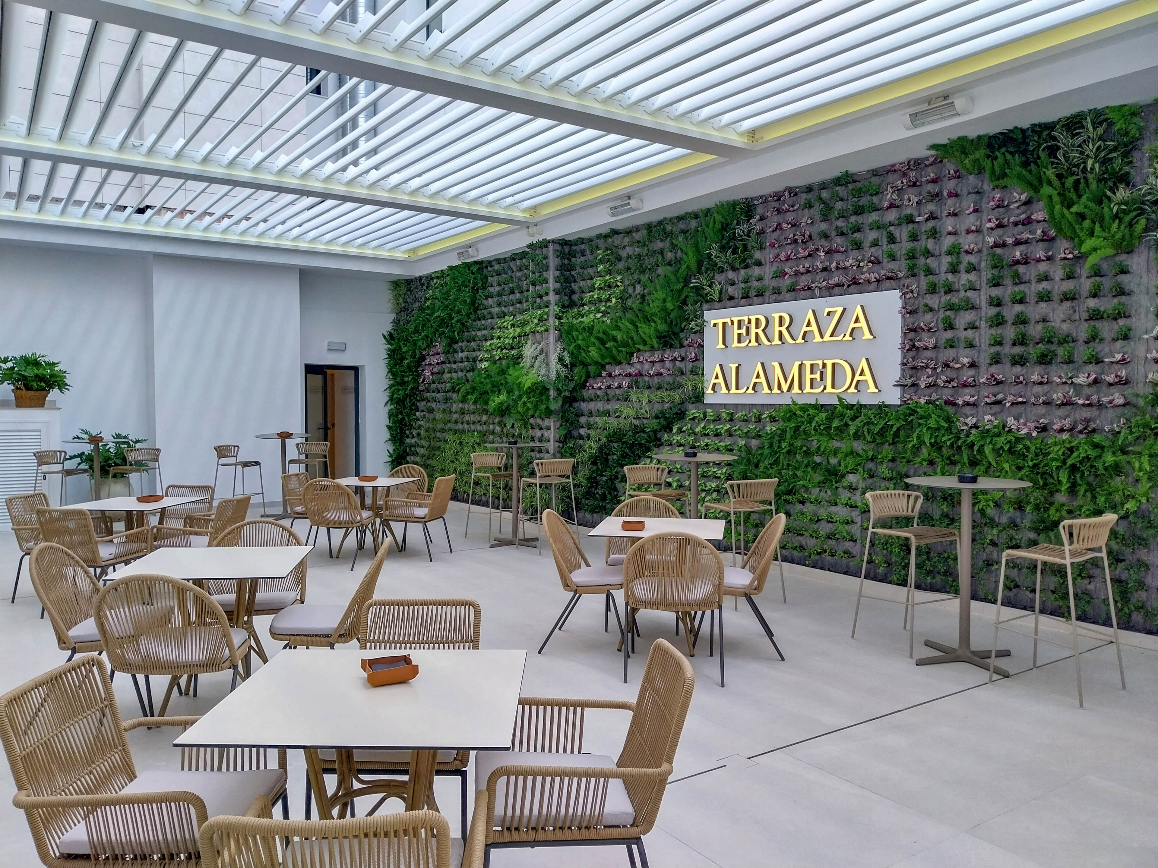 A contemporary terrace café features light wicker chairs and square tables arranged under a slatted glass roof. The focal point is a lush vertical garden wall with illuminated text reading “TERRAZA ALAMEDA.” Neutral flooring and minimalist décor create an airy, open atmosphere. The space appears ready for guests, with neatly set tables and abundant greenery providing a relaxed, modern ambiance.