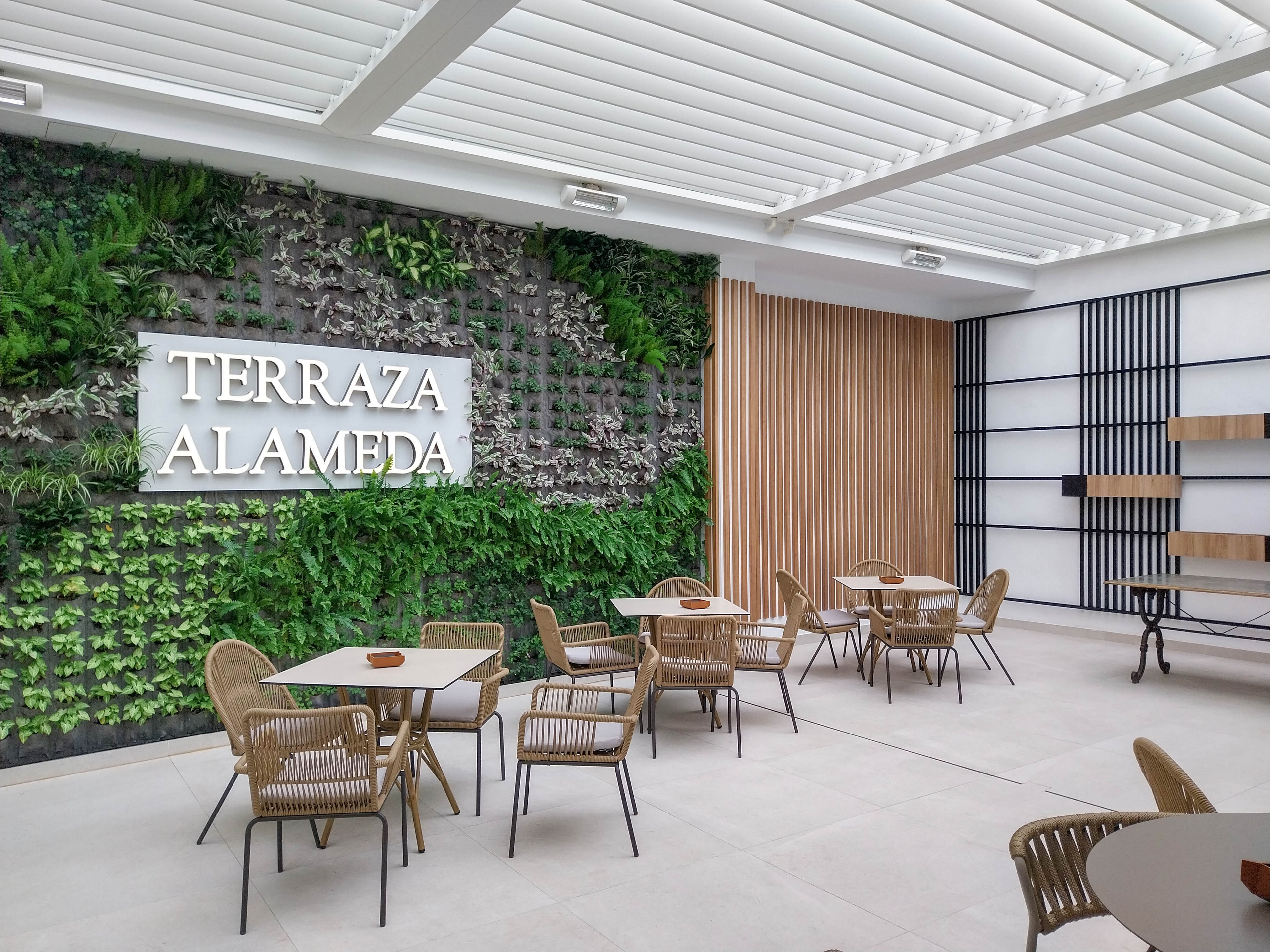 Contemporary terrace seating area labeled "TERRAZA ALAMEDA" features wicker chairs and round tables. A lush green vertical garden covers the wall behind the text, adding a natural focal point. The space is bright with a white slatted ceiling and light floor tiles, creating an airy atmosphere. Minimalist wooden slats and black shelving elements complete the modern design.
