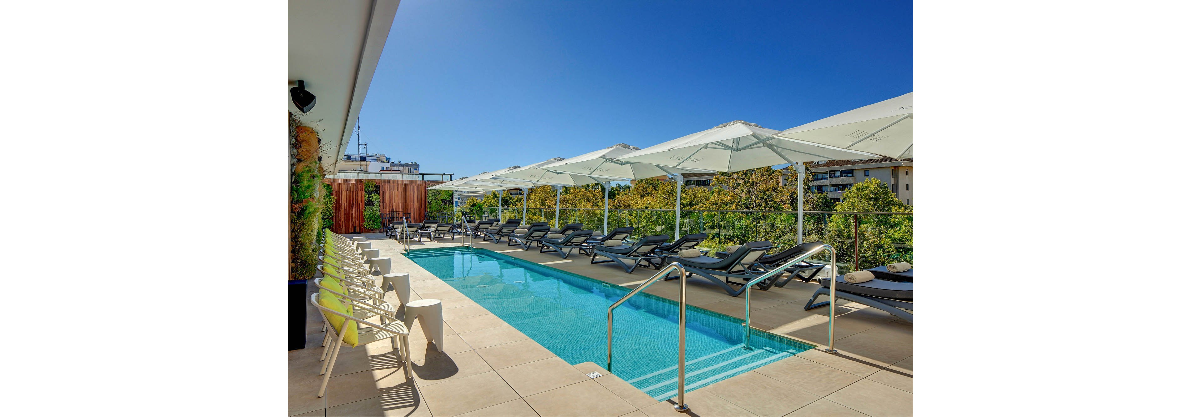 A modern rooftop swimming pool is lined with black sun loungers and large white umbrellas under a clear blue sky. The pool area features light-colored tiles, metal handrails, and a row of contemporary chairs and small side tables. Green trees and nearby buildings form the backdrop, creating an urban oasis atmosphere. The bright sunlight and clean design emphasize relaxation and leisure in an outdoor hotel or apartment setting.