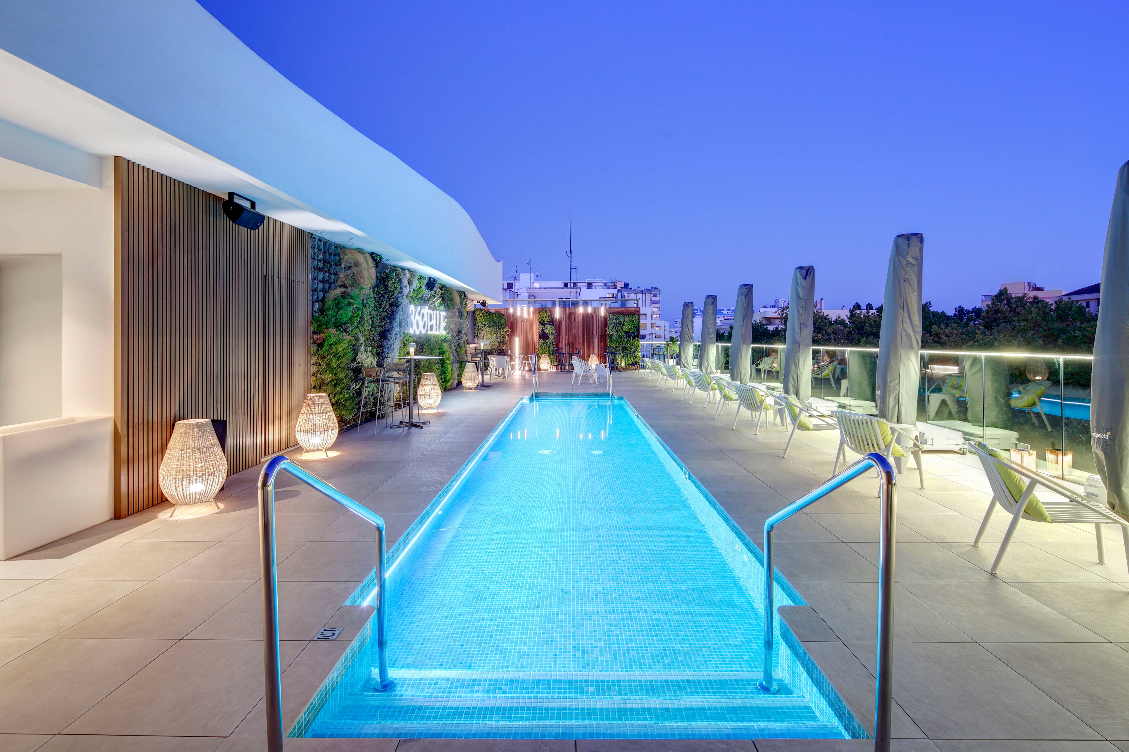 A sleek rooftop swimming pool stretches toward the city skyline under a clear evening sky. Contemporary lounge chairs and tables line both sides of the pool, creating a stylish relaxation area. Soft ambient lighting, including decorative floor lanterns, reflects on the water and surrounding surfaces. The scene conveys a luxurious, tranquil atmosphere ideal for leisure and nighttime socializing.