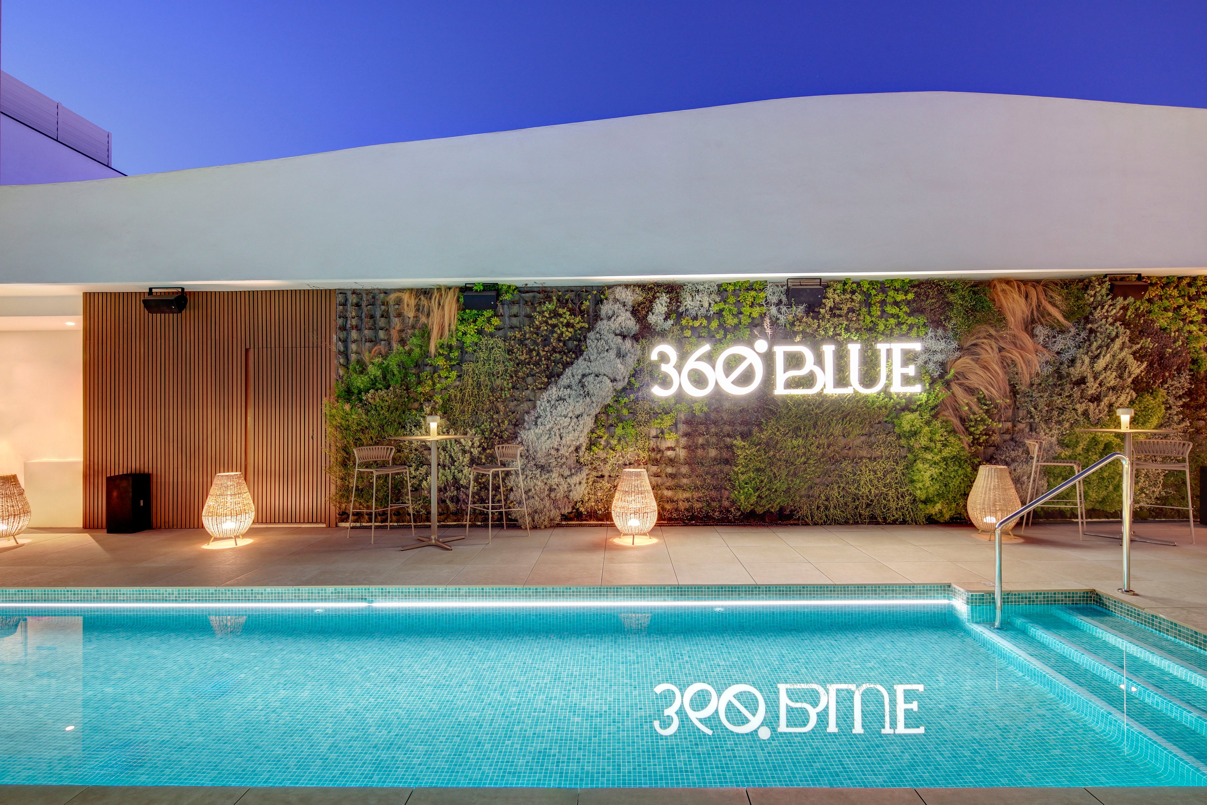A contemporary rooftop swimming pool is shown at dusk, reflecting the illuminated text '360 BLUE' on the water’s surface. The poolside area features minimalist railings, wicker lanterns, and small cocktail tables. Behind the pool, a vertical garden wall and wooden paneling add texture and greenery to the sleek architectural design. The tranquil evening sky enhances the calm, upscale atmosphere of the outdoor space.