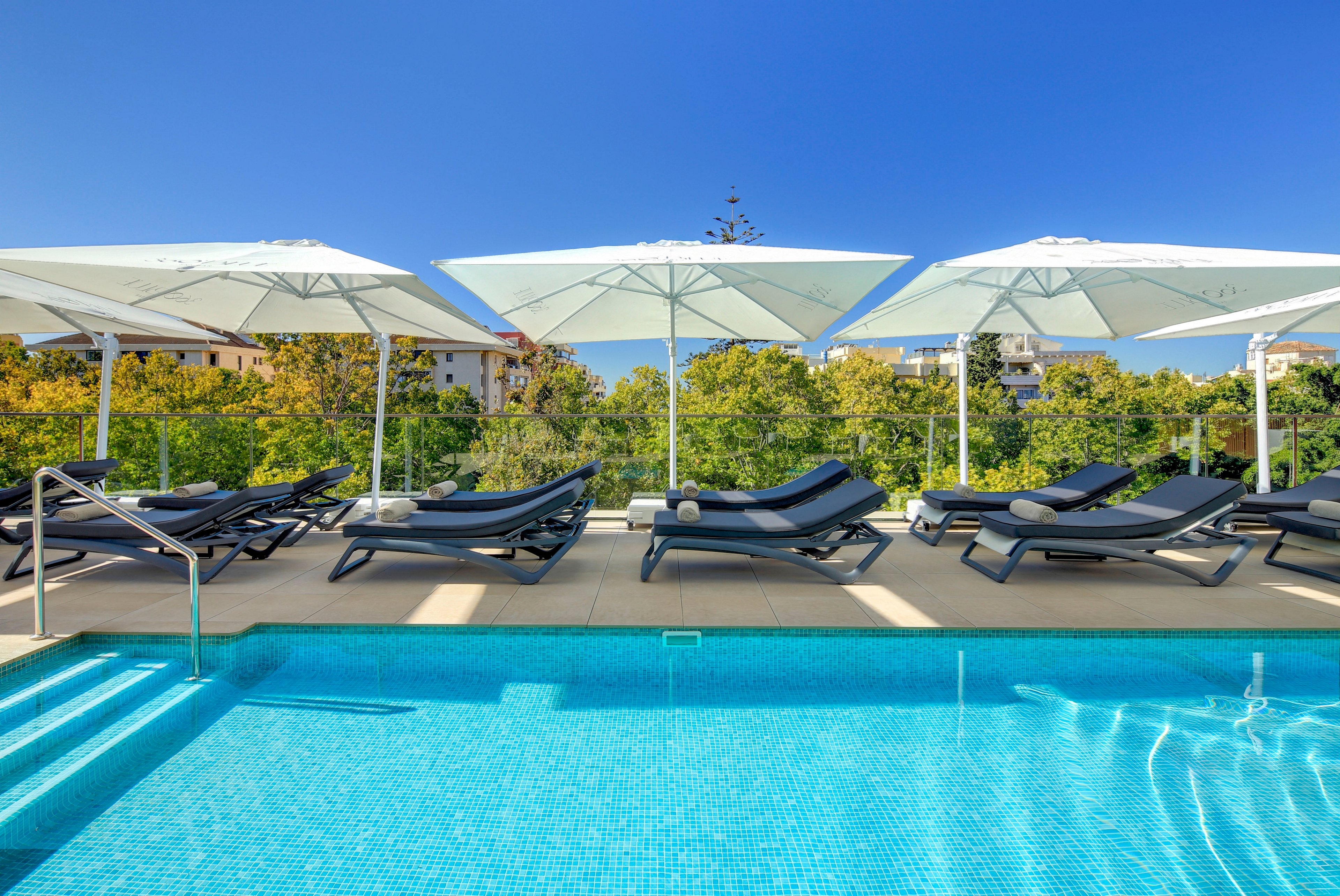 A bright outdoor swimming pool is surrounded by sleek black sun loungers and large white umbrellas. The clear blue water reflects the sunny sky, creating a refreshing resort atmosphere. Green trees and low residential buildings appear in the background beyond a glass barrier. The scene suggests a tranquil hotel or apartment terrace designed for relaxation and sunbathing.