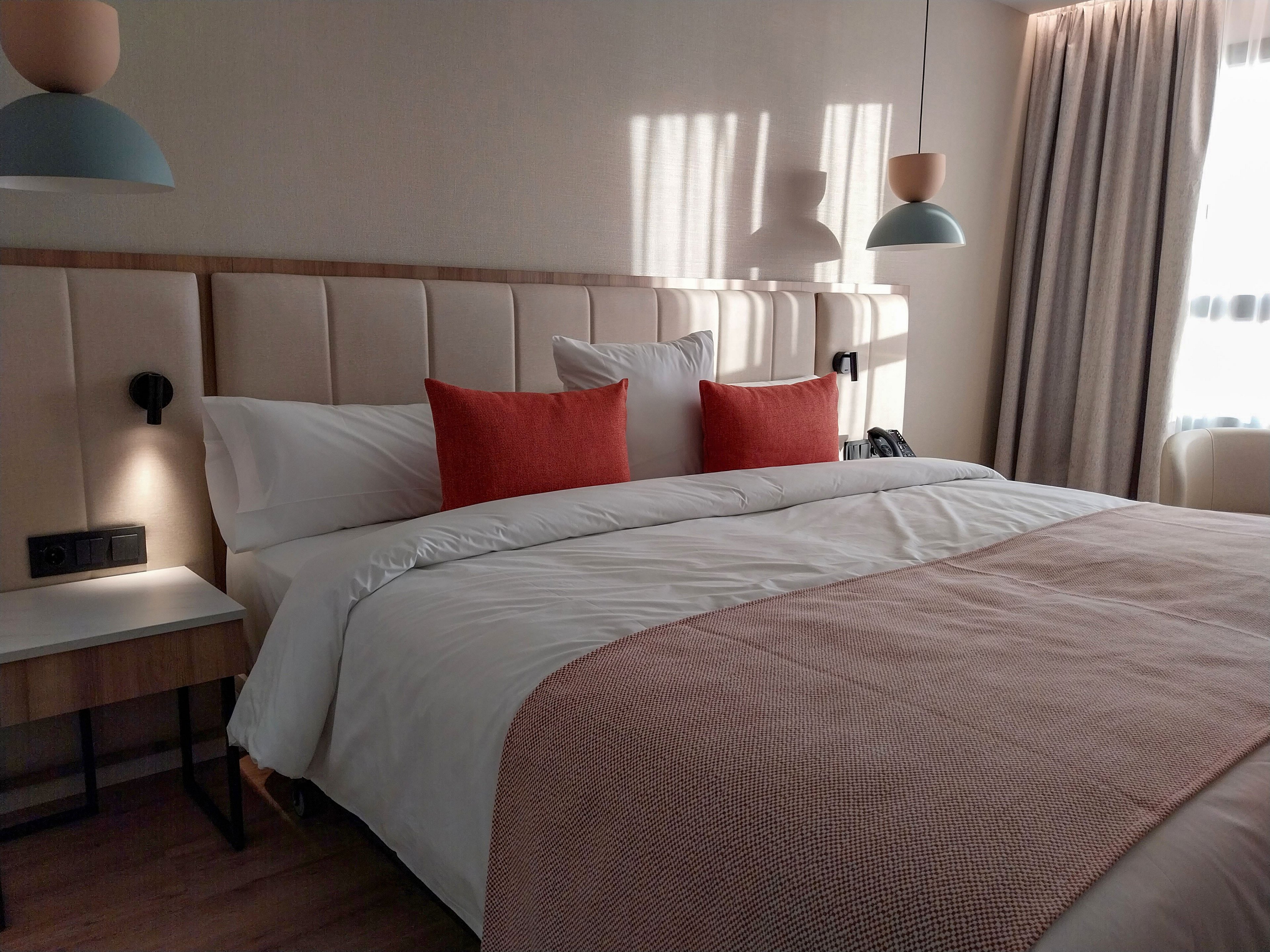 A neatly made double bed with white linens and a soft pink throw blanket is the focal point of this modern bedroom. Two coral accent pillows add a pop of color against the upholstered headboard. Sunlight filters through sheer curtains, casting soft shadows on the wall. Minimalist bedside tables, wall-mounted reading lights, and contemporary pendant lamps complete the cozy, stylish setting.