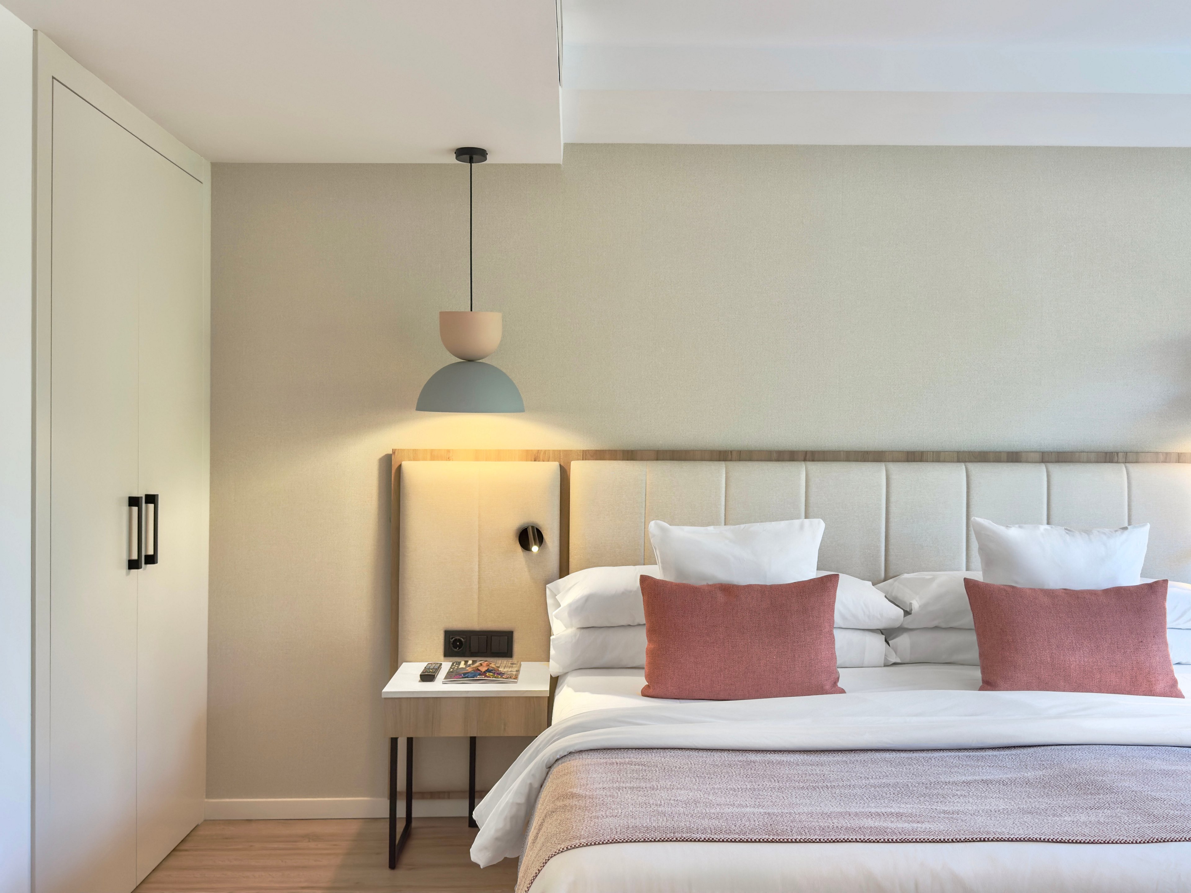 A contemporary bedroom features a neatly made bed with white linens and two muted pink accent pillows. The headboard is upholstered in a light fabric and framed by a wooden panel with integrated warm lighting. A small bedside table holds books and a phone, next to a wall-mounted reading light and power outlet. A modern pendant lamp hangs above the table, and a built-in wardrobe with double doors stands to the left, creating a clean, minimalist look.