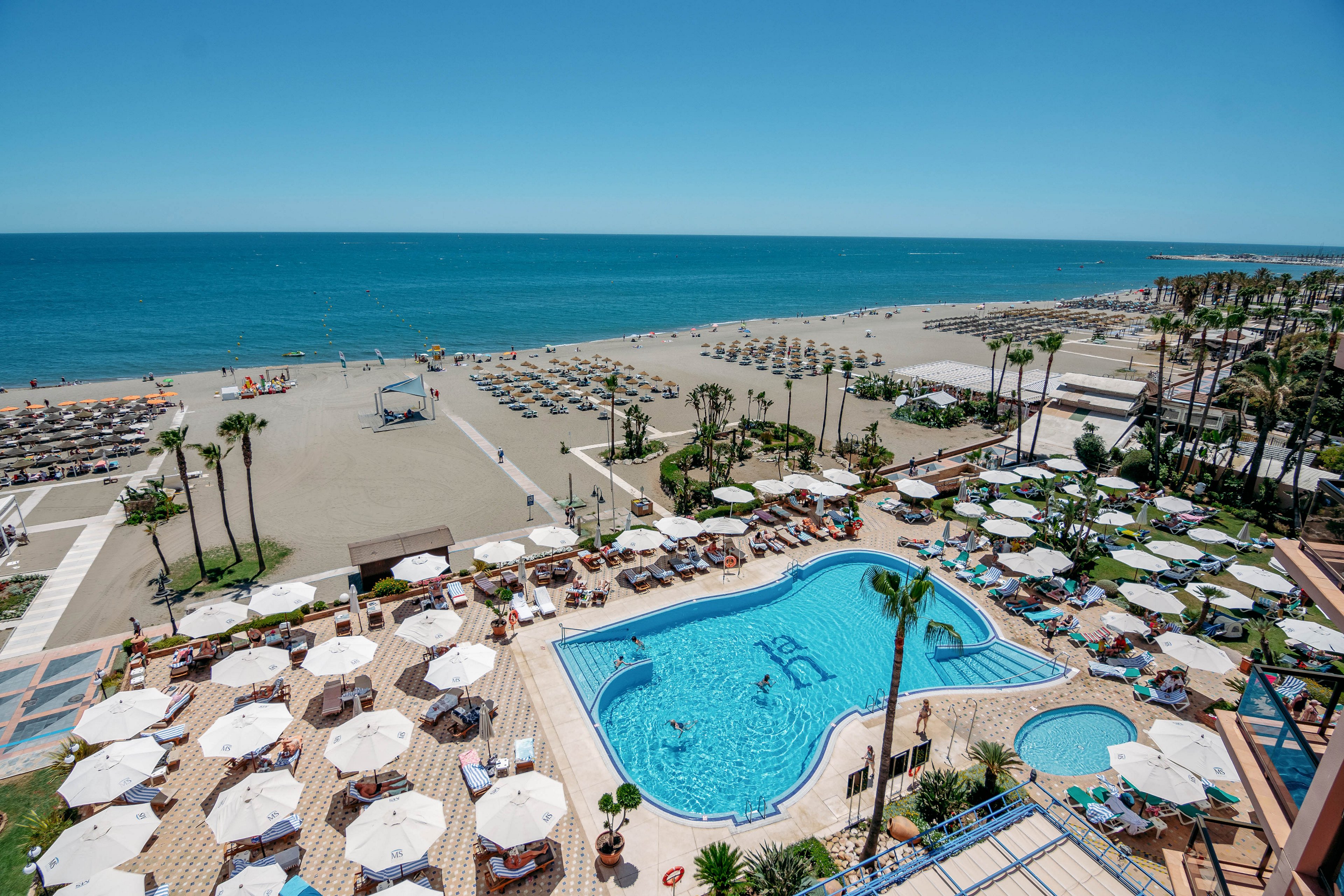 A large beachfront resort overlooks a bright blue outdoor swimming pool filled with guests. Numerous white sun umbrellas and loungers surround the pool area, creating a lively holiday atmosphere. Beyond the resort, a wide sandy beach stretches to the horizon, lined with organized rows of sunbeds and umbrellas. The calm sea and clear blue sky emphasize a sunny, relaxing coastal vacation setting.
